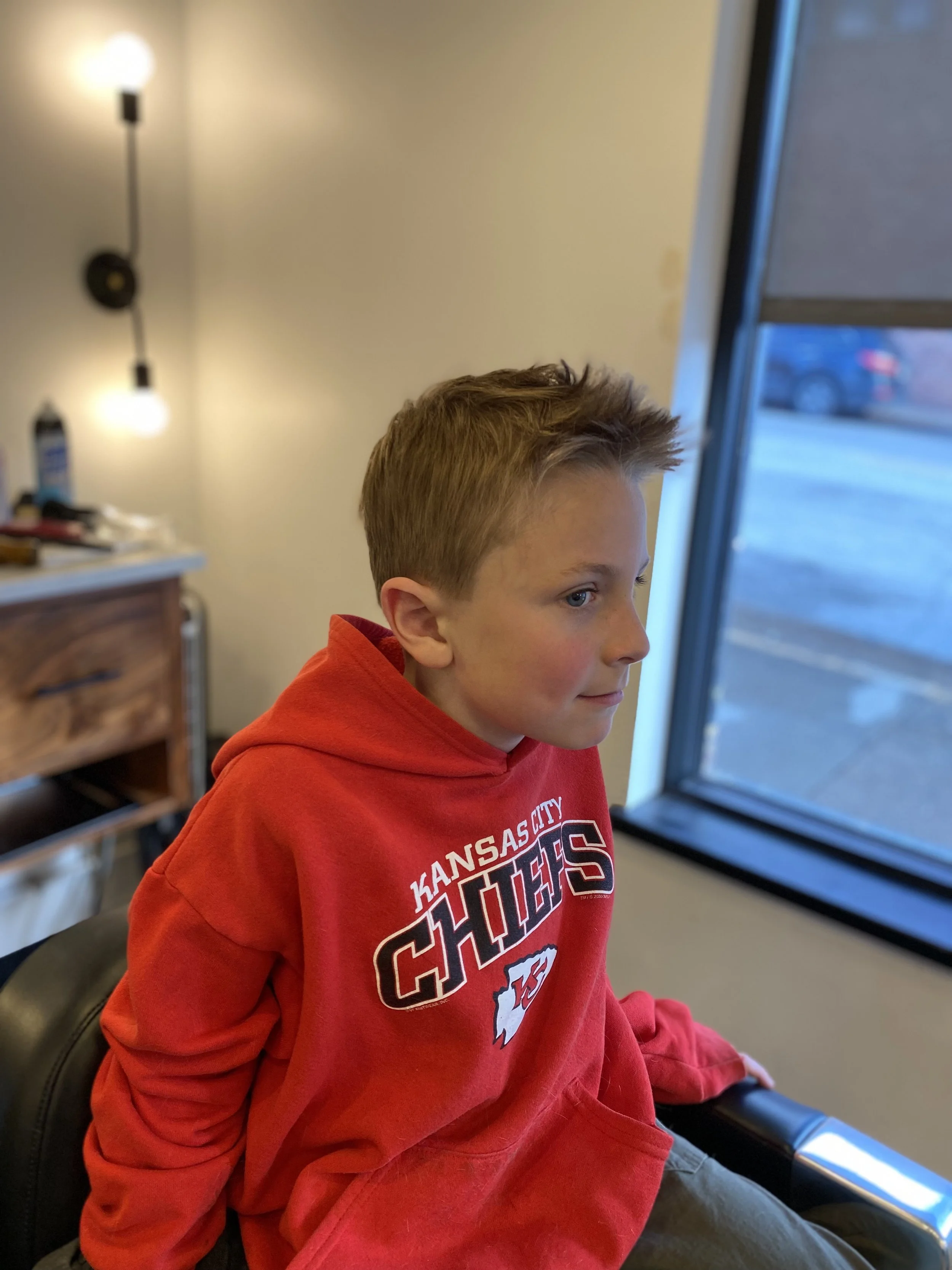 A young boy with short, light brown hair and blue eyes, wearing a red Kansas City Chiefs hoodie, sitting indoors near a window and a wall with a light fixture.