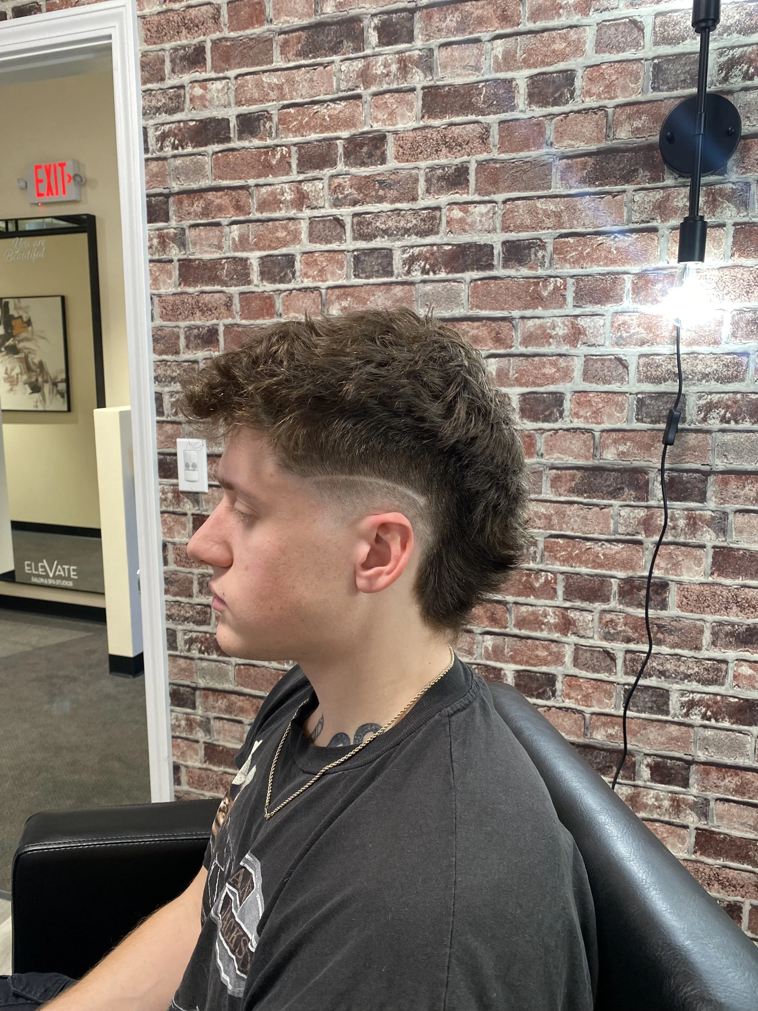 A young man with brown hair and a fade haircut sitting in a barber shop chair against an exposed red brick wall, with a black leather apron and a black T-shirt.