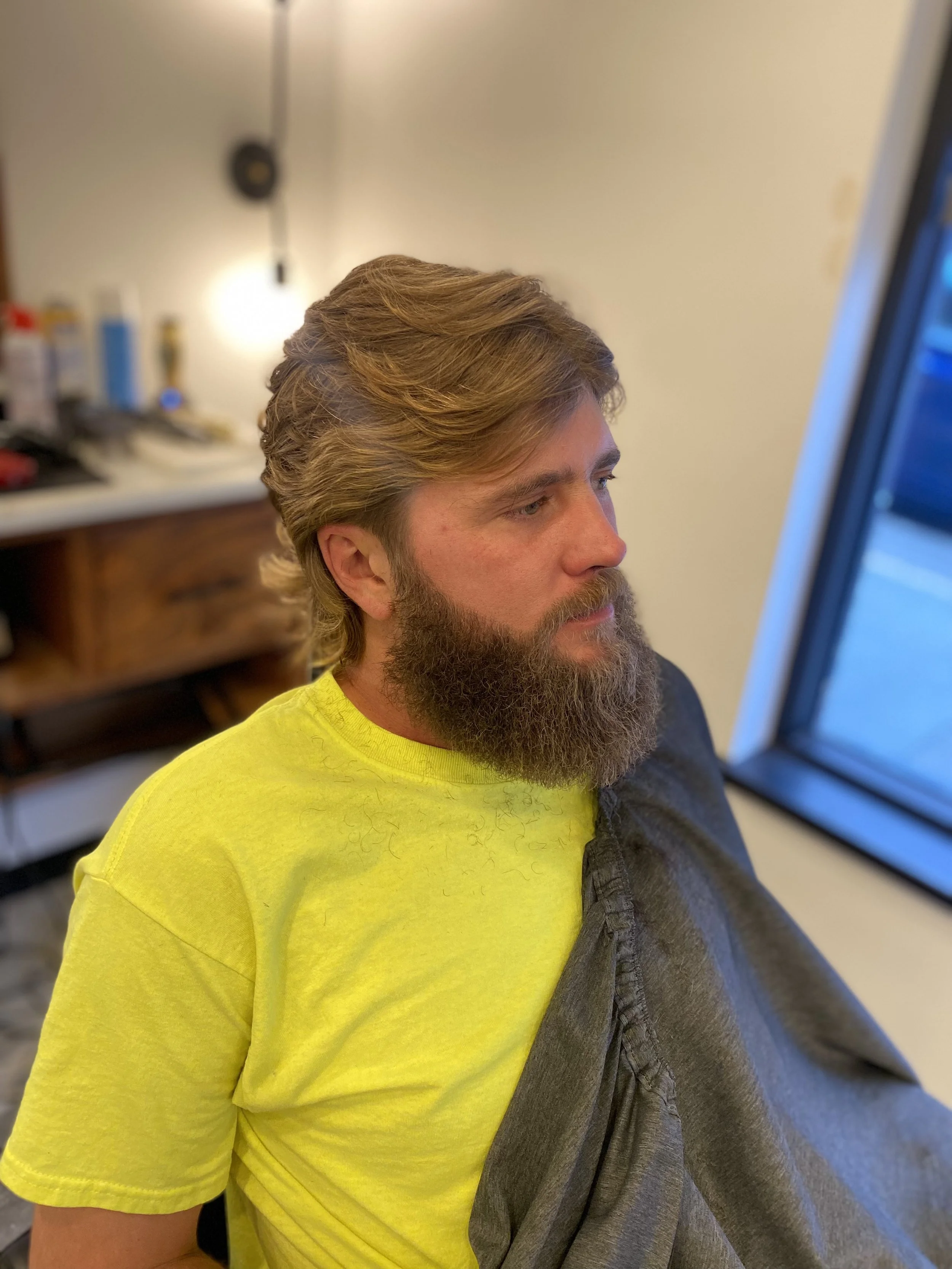 A man with a full beard and long, wavy, light brown hair is sitting indoors, looking to his right at a screen. He is wearing a bright yellow T-shirt and a gray jacket. The background includes a blurry white wall, some shelves with bottles, and a part