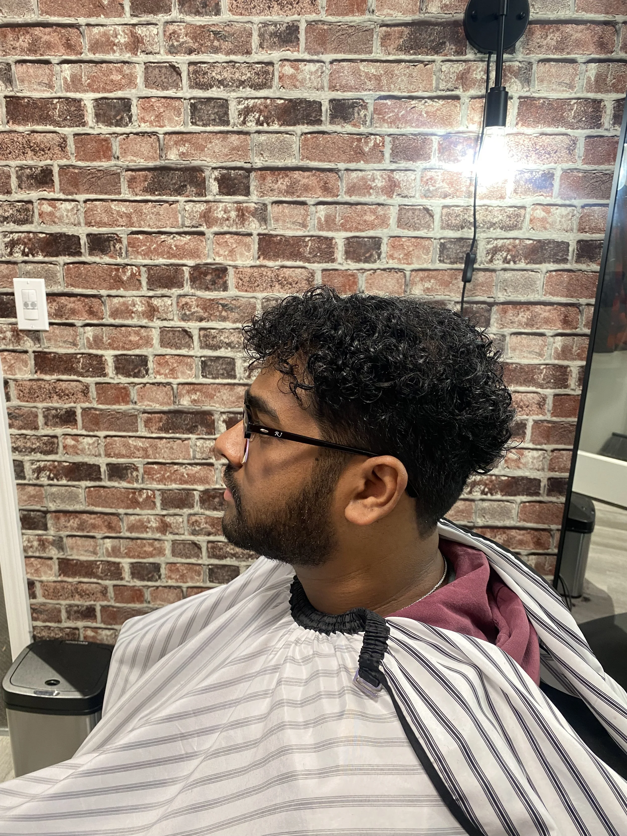 A man with dark, curly hair and glasses getting a haircut in a salon with a brick wall background.
