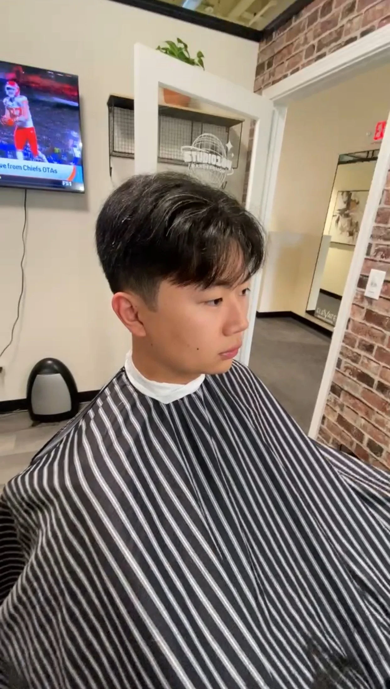 Young man getting a haircut at a salon, wearing a striped barber cape, and sitting in front of a mirror.