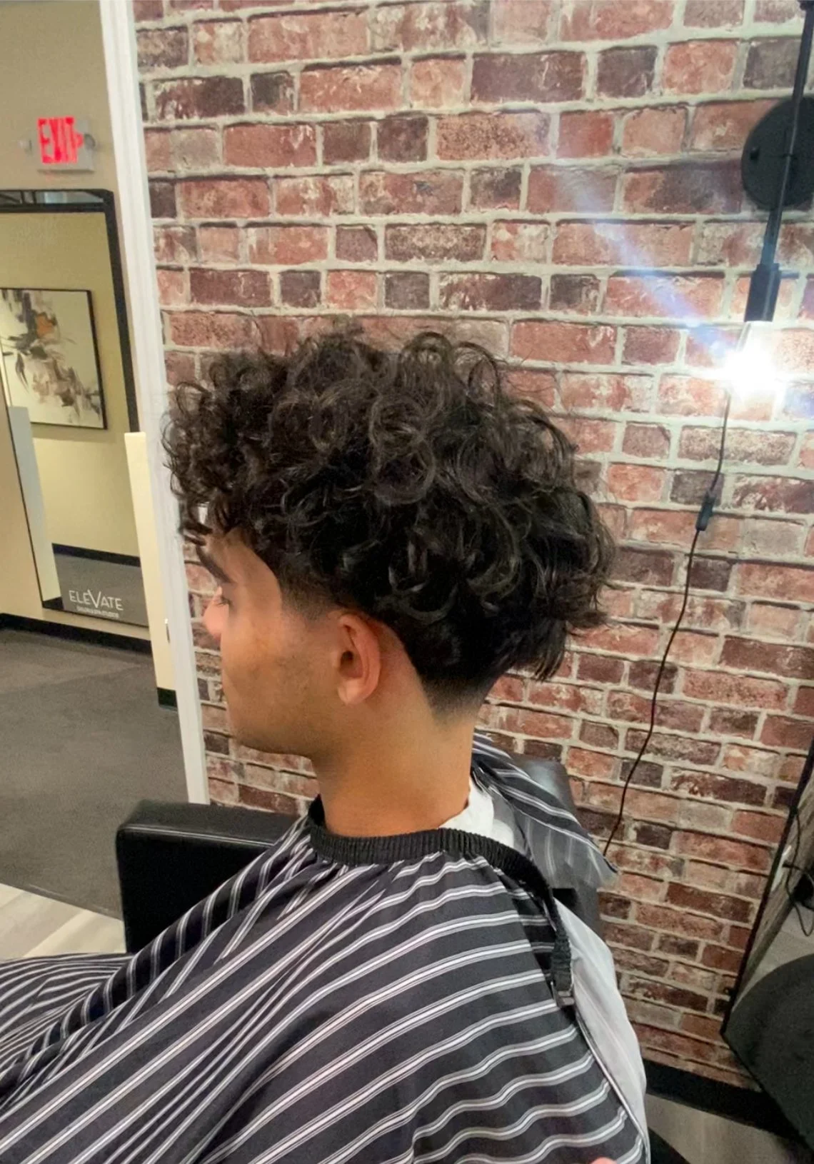 Young man with curly dark hair getting a haircut at a salon, seated in front of a brick wall, with a barber cape with white and black stripes.