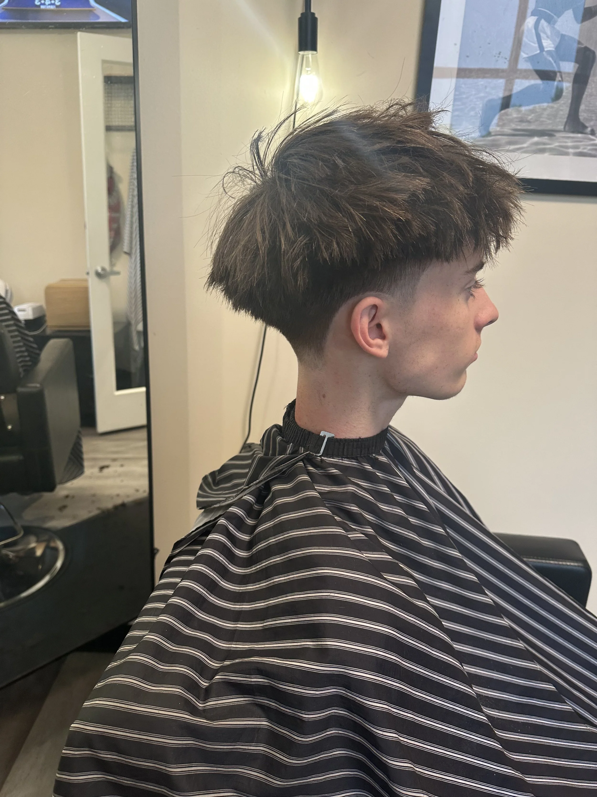 A young man with styled short hair sitting in a barber's chair, wearing a black and white striped barber cape, in a barbershop with a mirror and framed picture on the wall.
