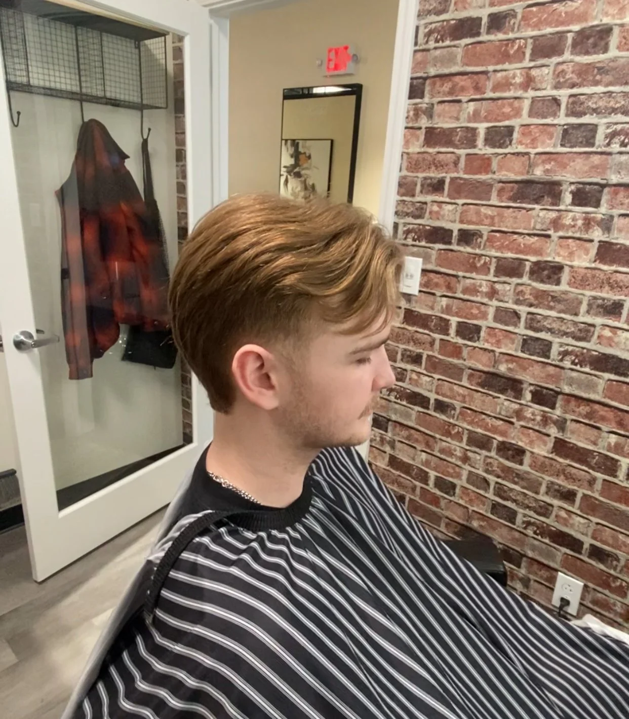 Young man with styled brown hair sitting in a barber's chair, wearing a black and white striped cape, in a barbershop with brick wall decor.