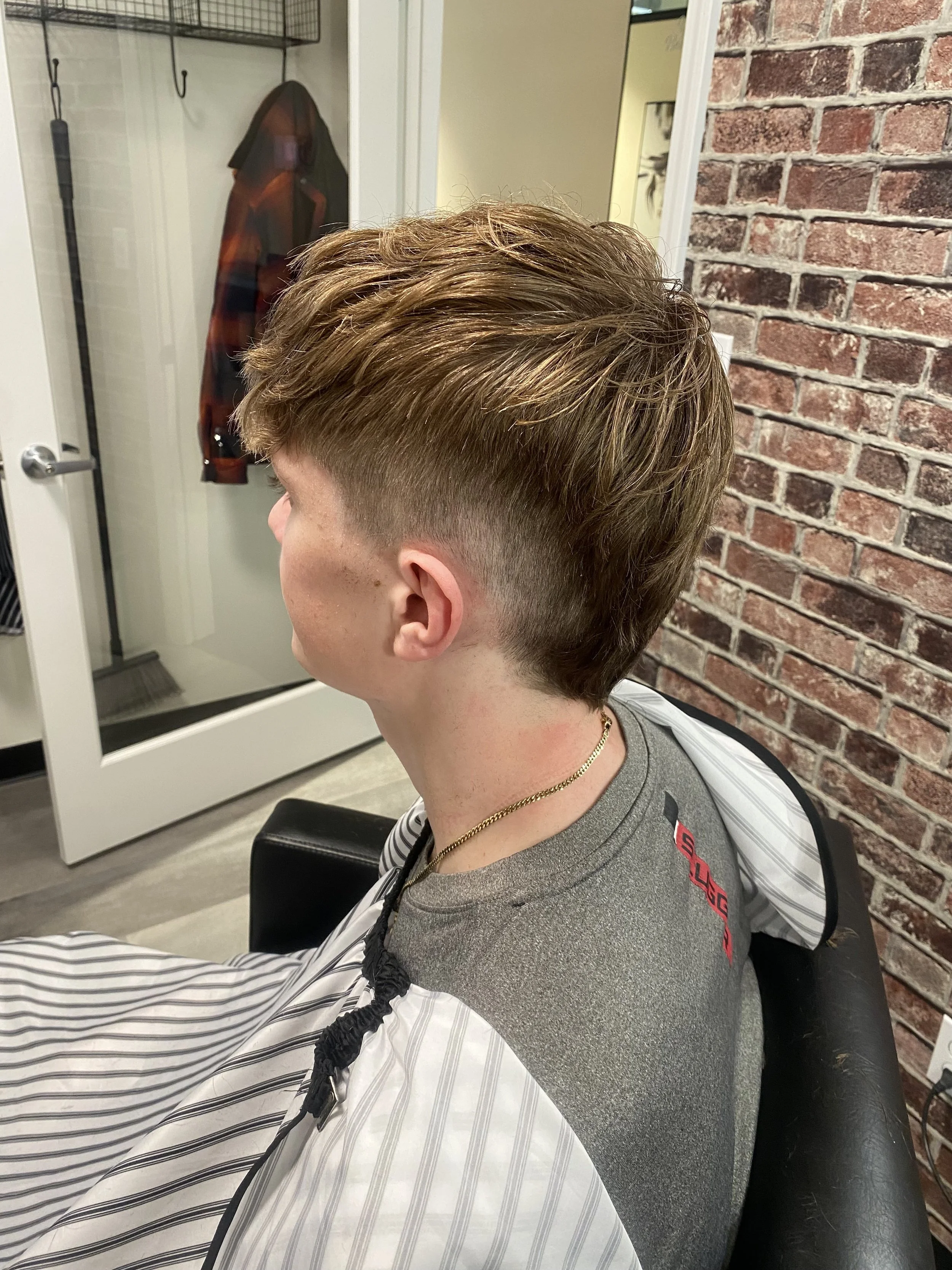 A young person with a short, styled haircut sitting in a barber shop chair. The background shows a brick wall and a door with hooks and a coat hanging on it.