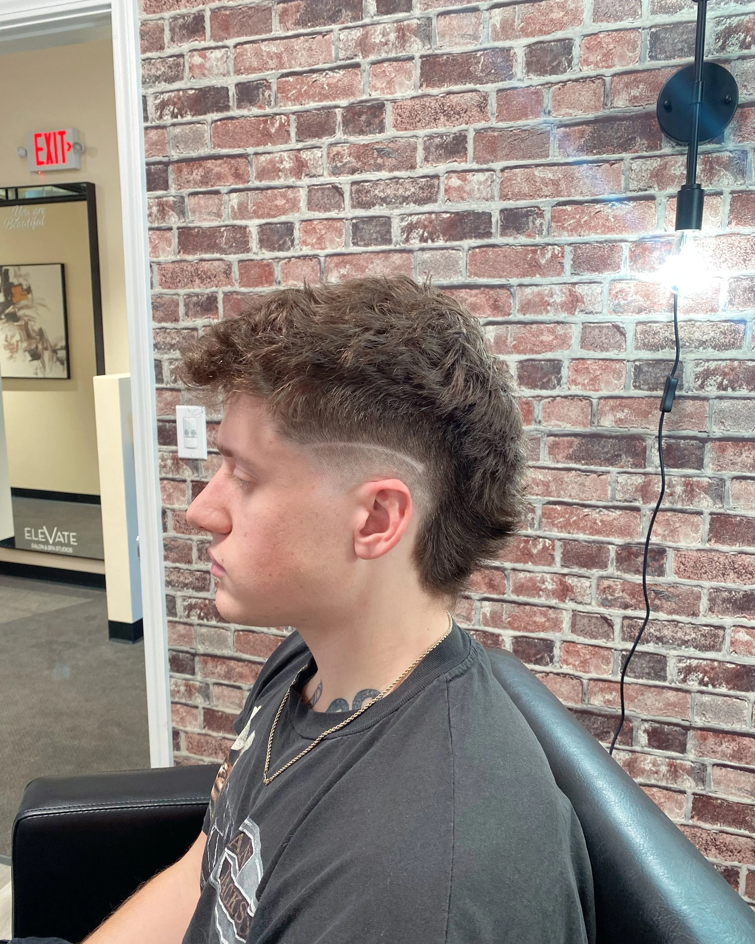 Profile view of a young man with short, curly hair, sitting in a salon or barbershop chair. The background features a red brick wall, a black wall-mounted lamp, and a decorative mirror with a sign that says "You are Beautiful." The man is wearing a b