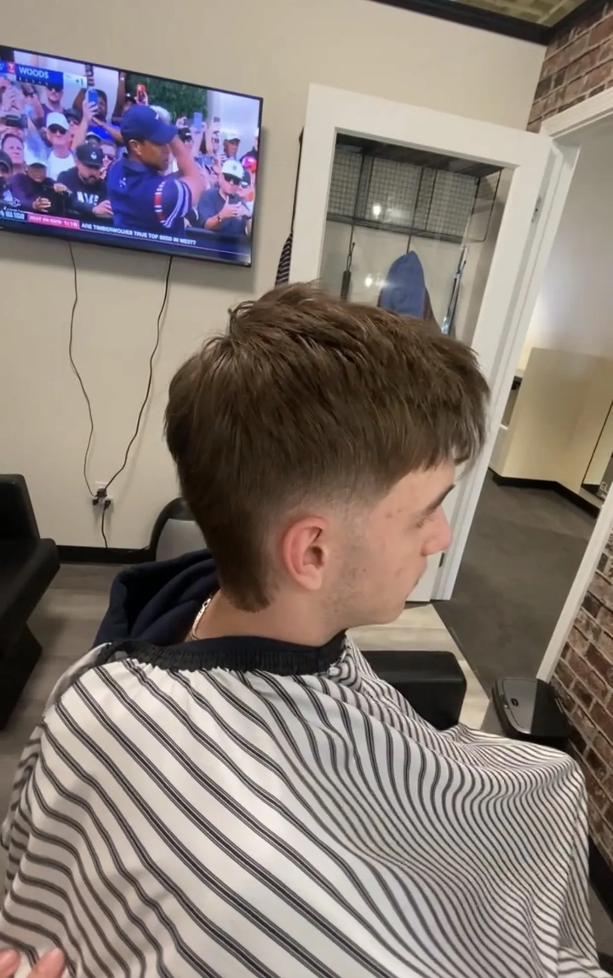 A young man with brown hair and light skin sitting in a barber's chair, wearing a striped barber cape, looking downward, inside a barbershop with a brick wall and a TV screen showing a golf tournament in the background.