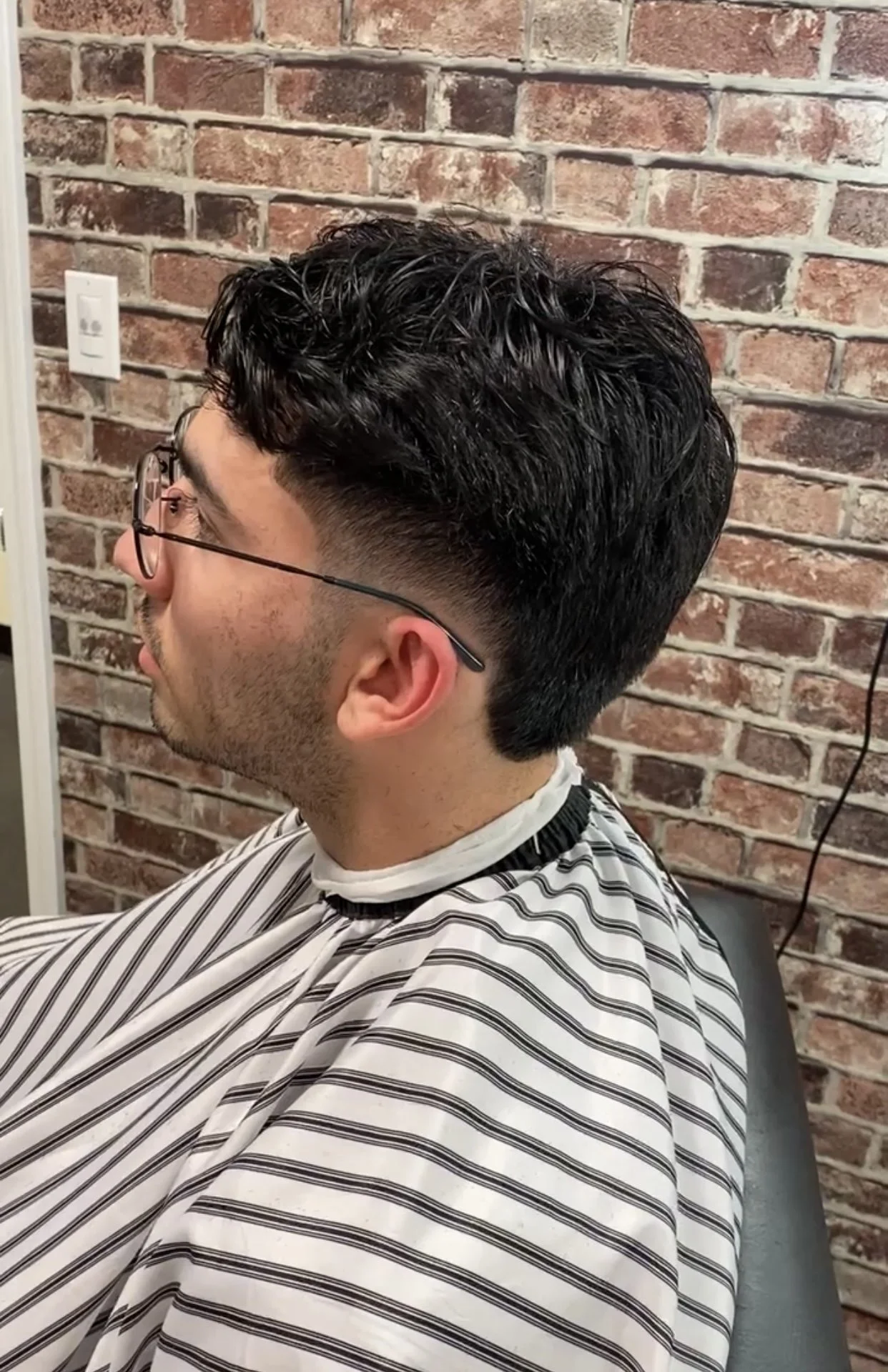 A man with dark, curly hair and glasses getting a haircut in a barbershop with a brick wall background.