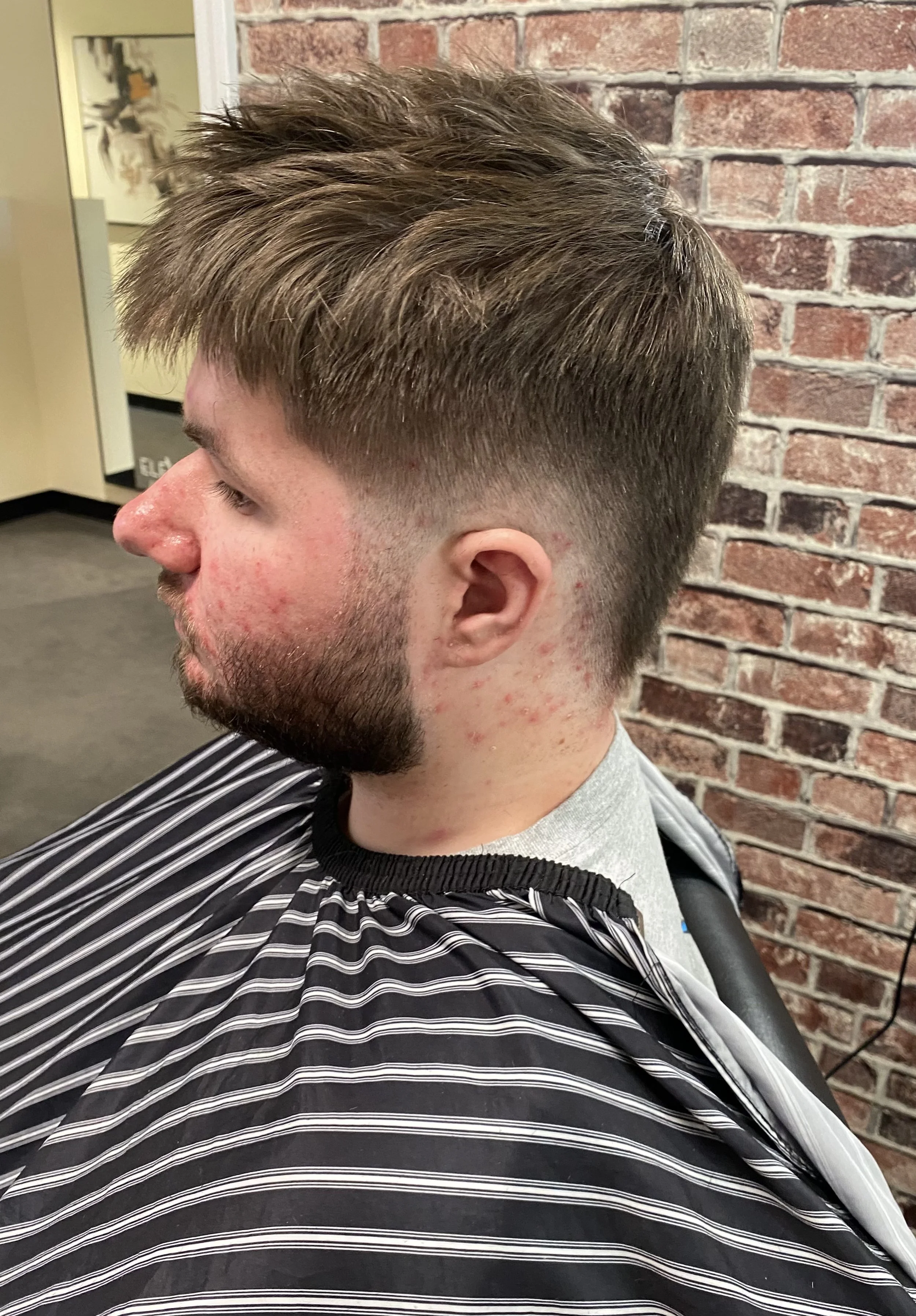 Side profile of a man with a short, layered haircut at a barbershop. He has some red skin and a beard, sitting in a chair with a striped barber cape. The background includes a brick wall and some framed art.