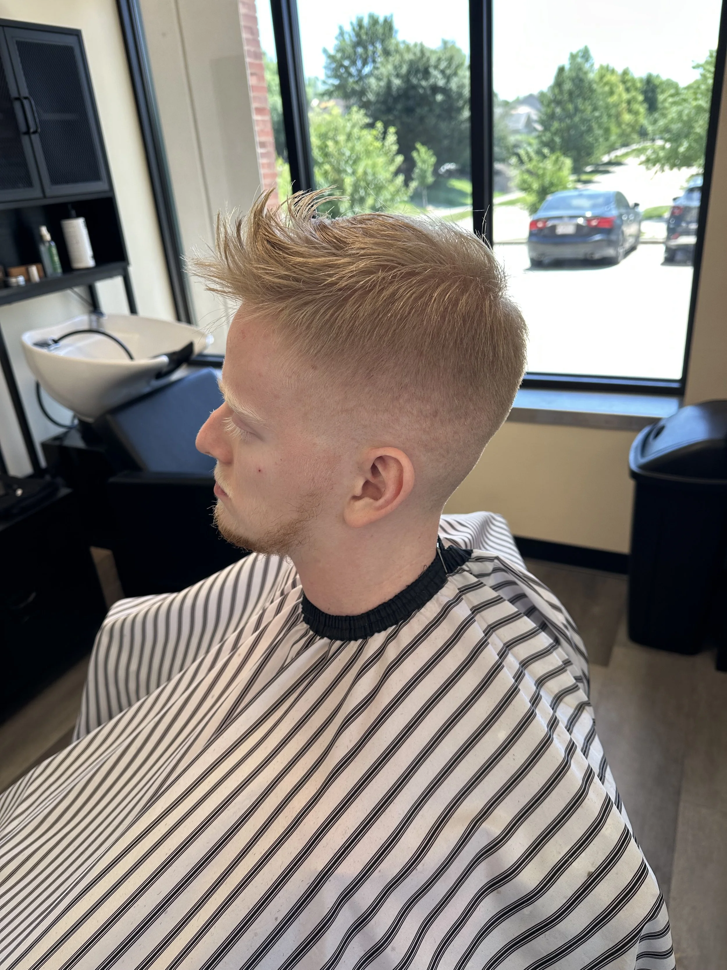 A person with blondish hair styled in a spiked manner getting a haircut in a salon, sitting in front of a large window showing a parking lot and green trees outside.
