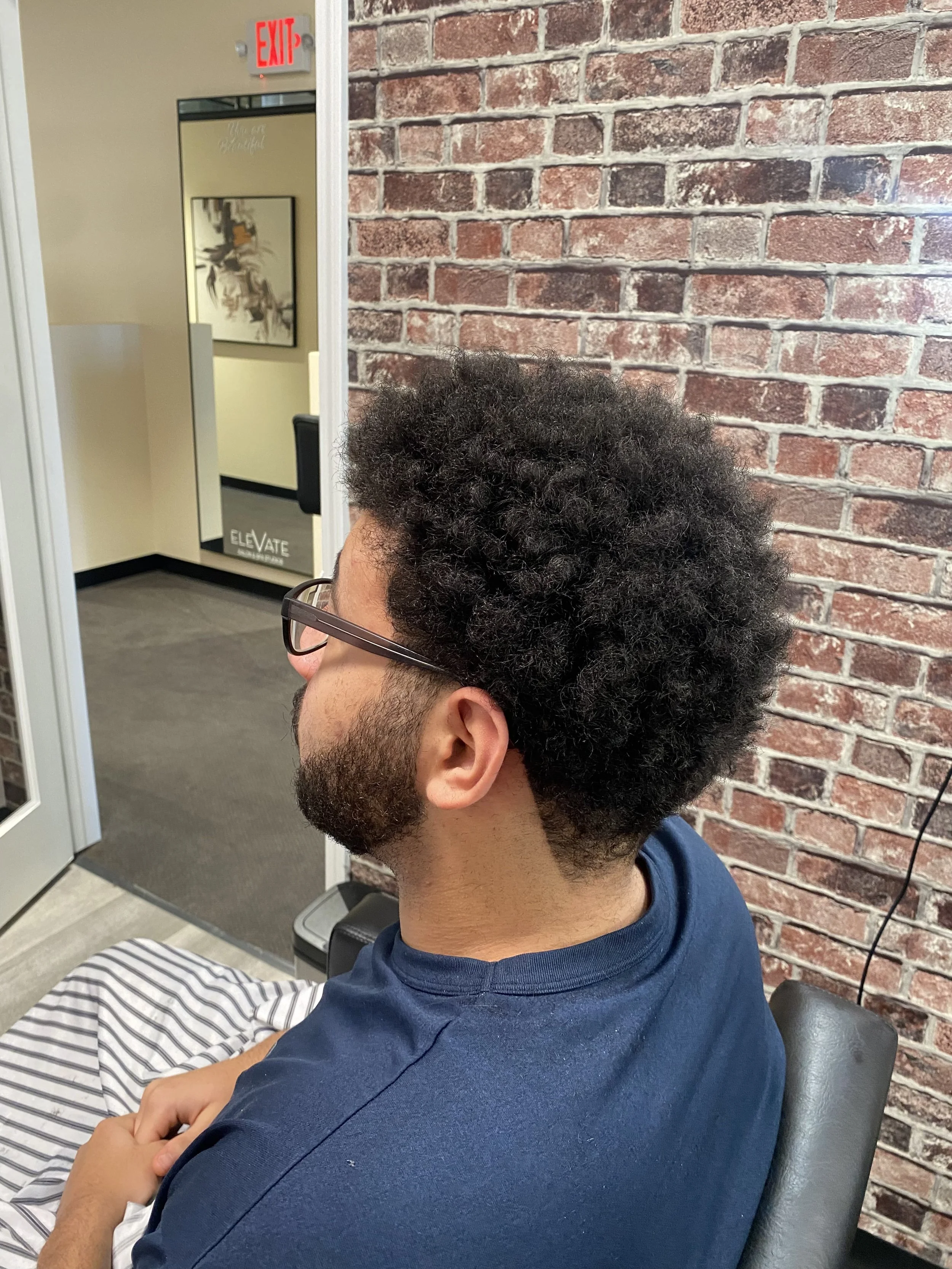 A man with curly dark hair and glasses sitting in a chair at a salon or barbershop, with a brick wall in the background.