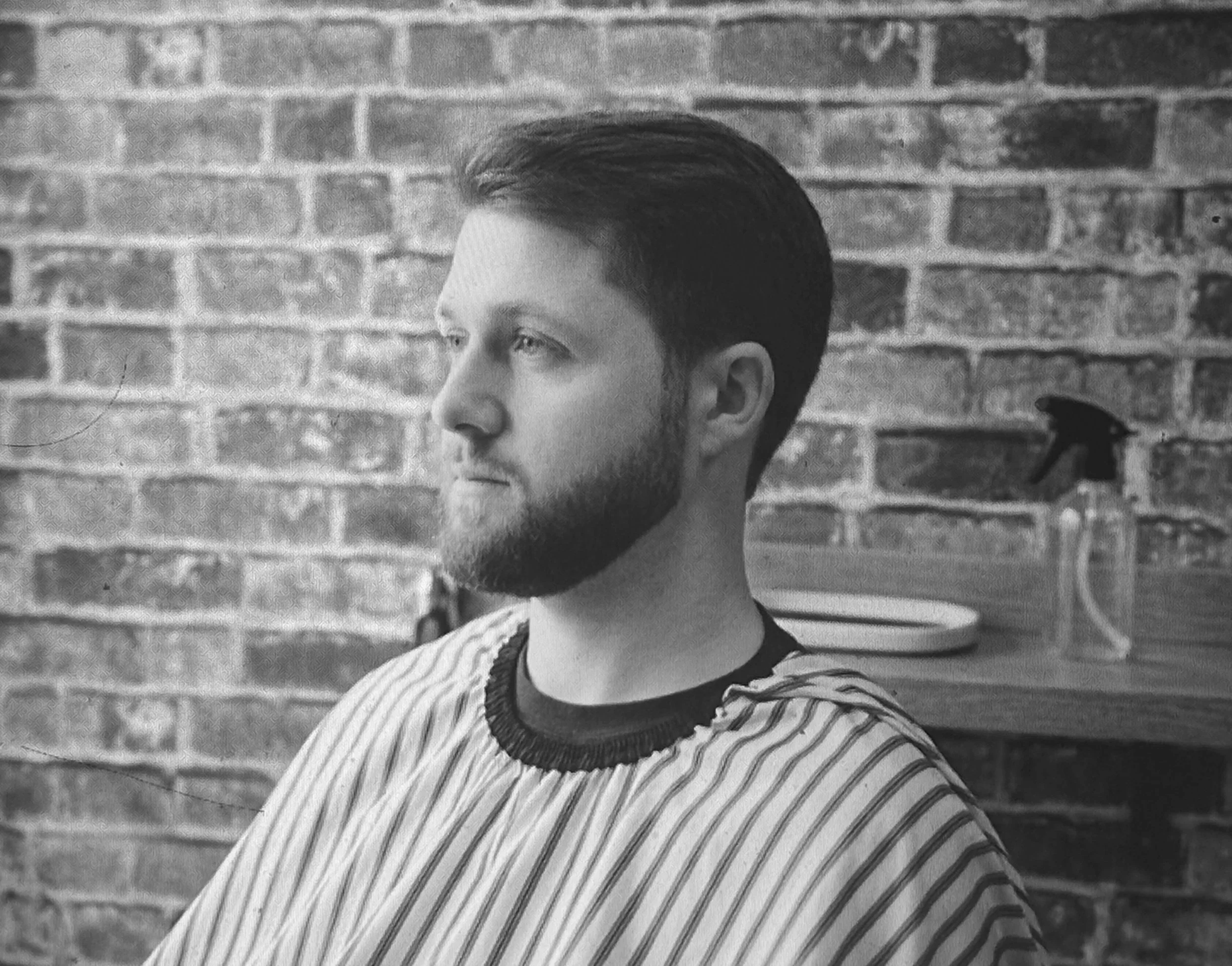 Black and white photo of a young man with short dark hair and a beard, wearing a striped shirt, standing in front of an exposed brick wall.