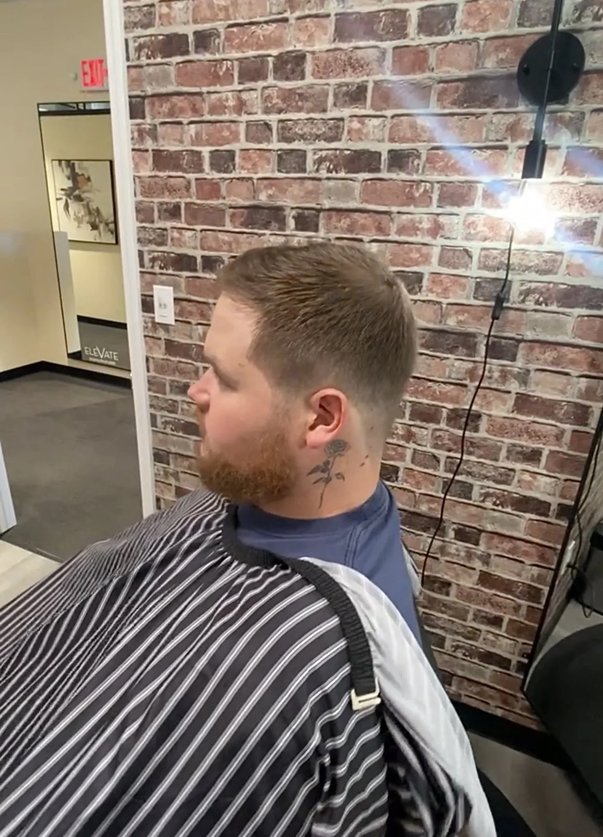 A young man with a beard and a tattoo on his neck is sitting in a barber shop chair getting a new haircut. He is wearing a cape with black and white stripes, and the background features a brick wall with a wall-mounted light.
