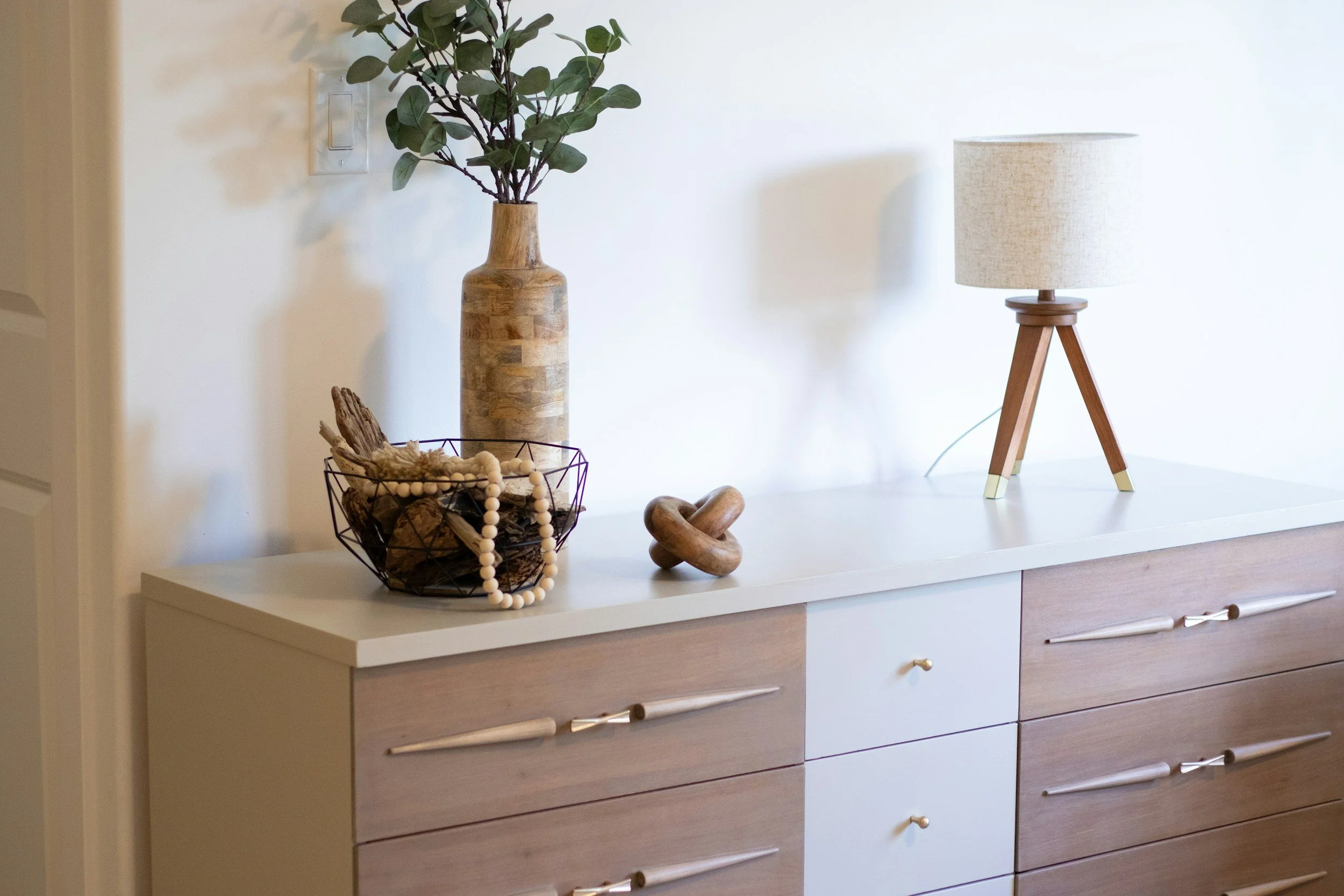 A modern dresser with a white top and wooden drawers, decorated with a tall wooden vase with green leafy branches, a wire basket with decorative wood pieces and a string of beads, and a wooden tripod table with a beige lampshade.