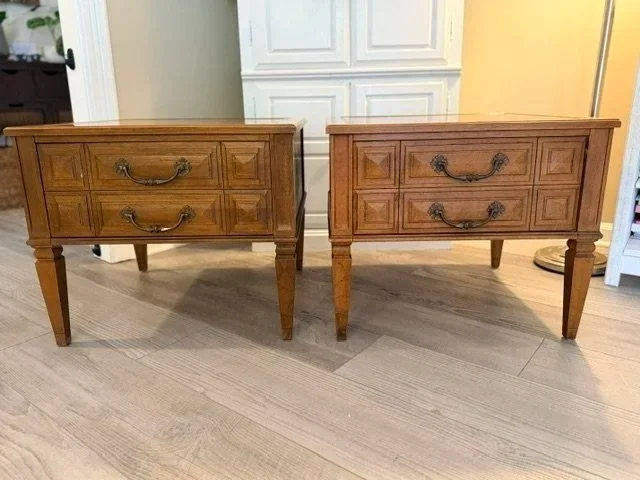 Two wooden nightstands with drawers and curved handles, standing on tapered legs, in a room with light-colored wood flooring and a white paneled door in the background.