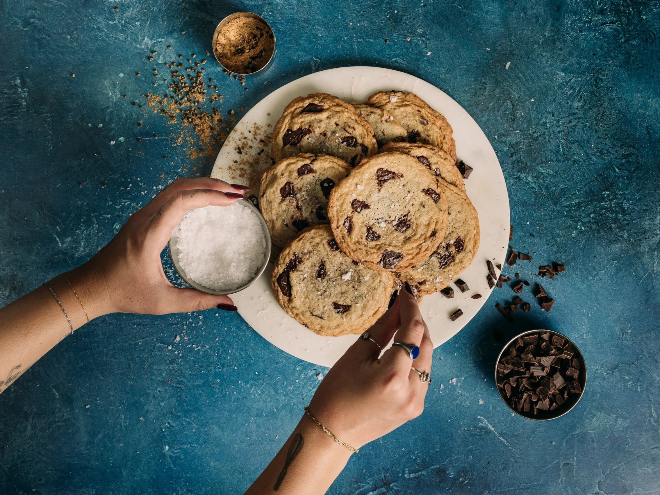Overhead view of chocolate chip cookies with sea salt on a white plate, surrounded by baking ingredients and hands on a blue table