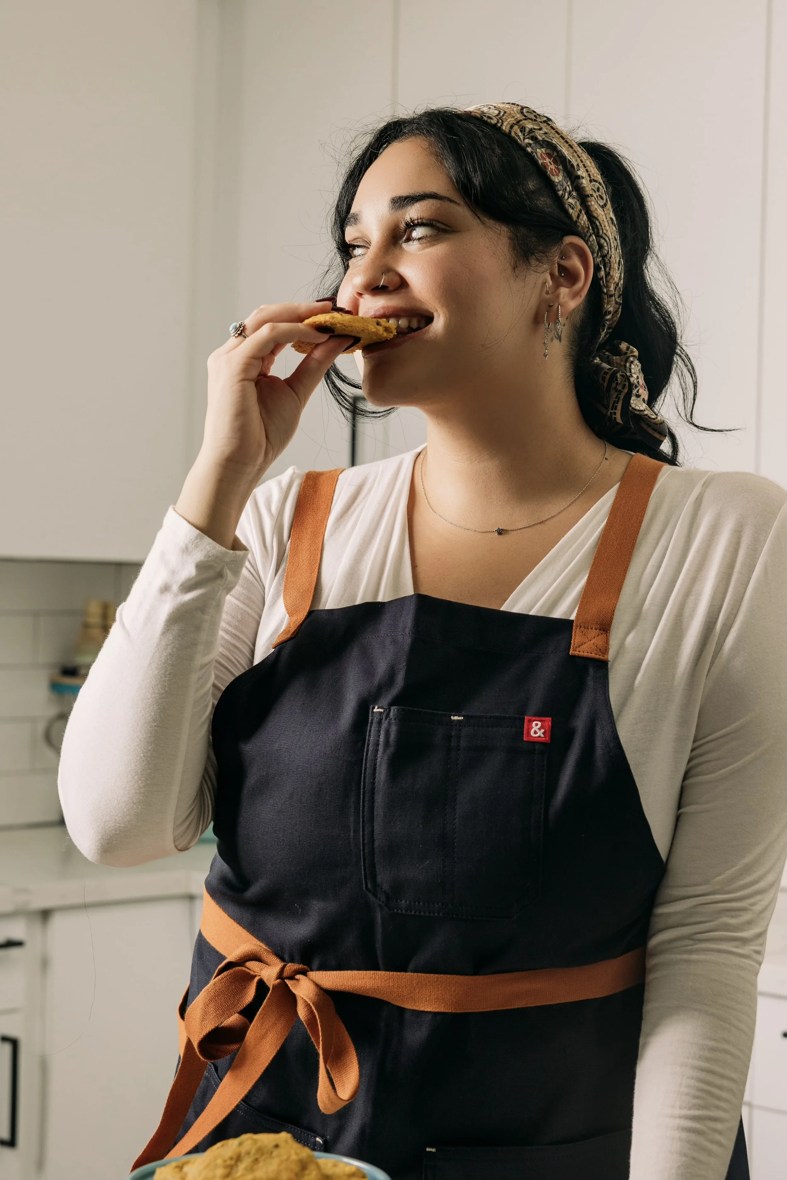 Woman wearing an apron and patterned headband takes a bite of a cookie in a kitchen.