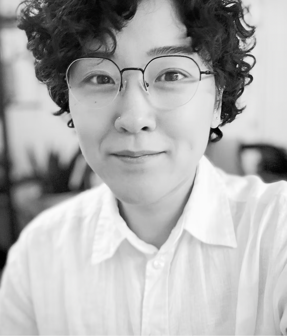 Close-up black and white portrait of a young person with curly hair, wearing glasses and a nose piercing, smiling slightly, dressed in a white collared shirt, with a blurred background.