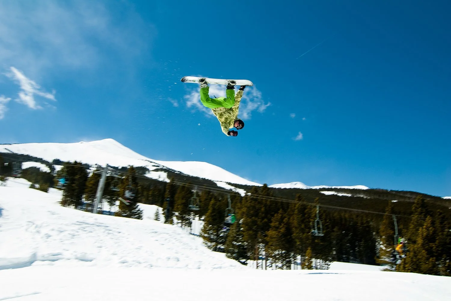 A skier in a green jacket and camouflage pants performing a backflip in mid-air on a snowy mountain slope under a clear blue sky, with ski lift chairs and snow-covered trees in the background.