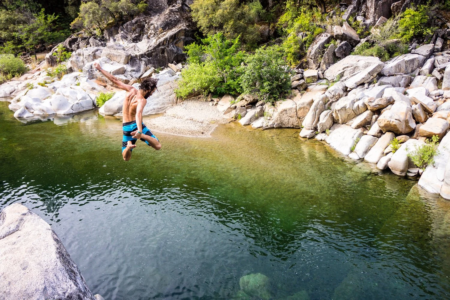A man jumping into a river from a large rock in a natural outdoor setting with rocks and trees.