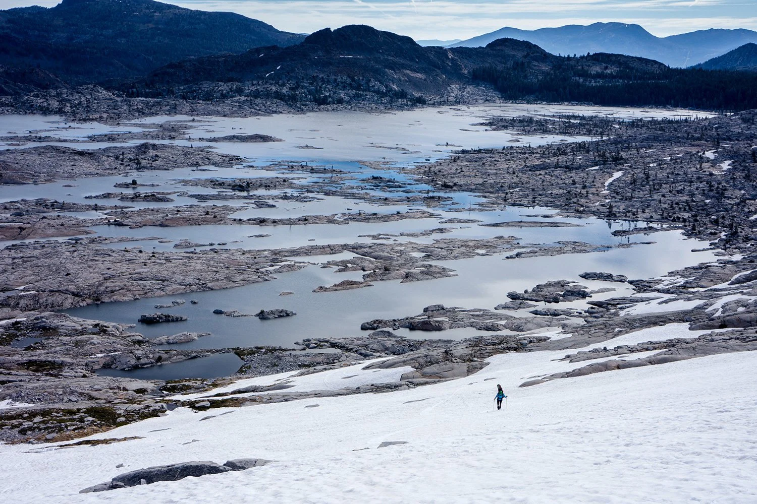 A person hiking on snow-covered terrain overlooking a lake, rocky landscape, and distant mountains under a cloudy sky.
