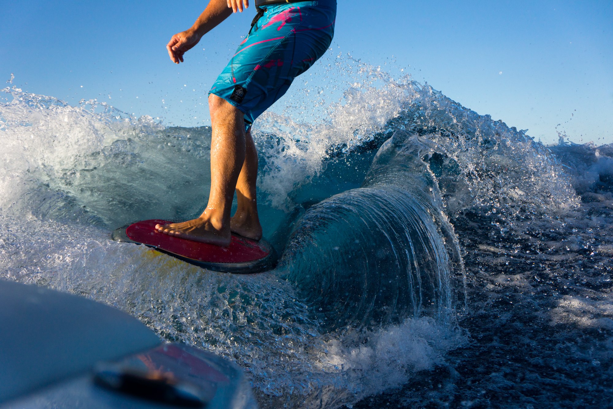 A person surfing on a wave, wearing blue and pink swim trunks, standing on a red surfboard with a pink nose, with water splashing around them under a clear blue sky.