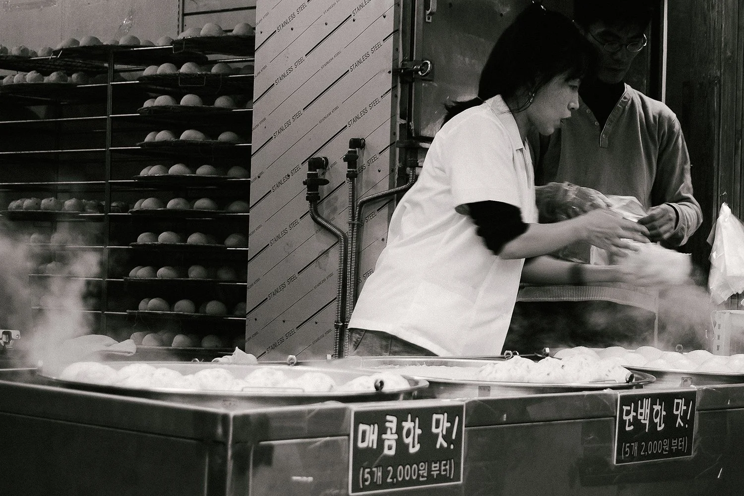Two people working at a food stall with steam rising from cooked food, metal racks with round items in the background, and Korean signs on the front of the stall.