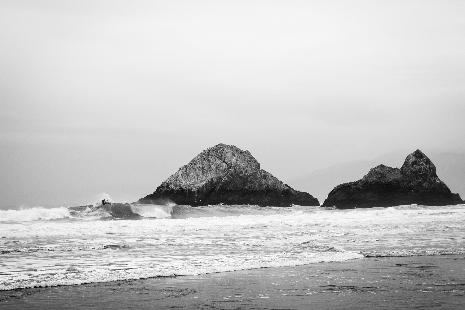Black and white photograph of two large rocks protruding from the ocean near the shoreline, with a person surfing a wave in the water.