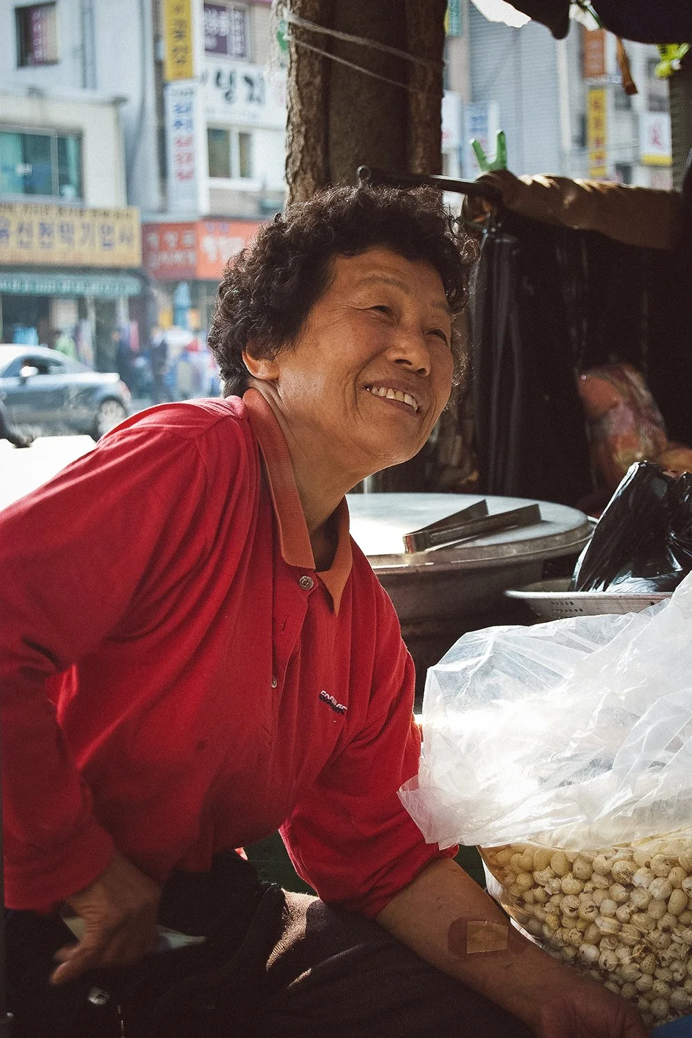 Smiling woman in red shirt selling packaged snacks at an outdoor market with cars and buildings in the background.