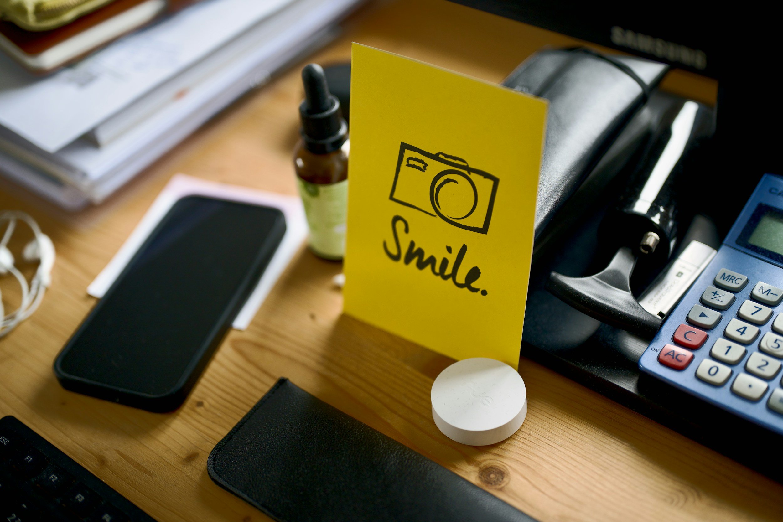 A cluttered desk with a yellow sign that says 'smile' and a drawing of a camera, along with various office supplies including a phone, calculator, and a small bottle of essential oil or medication.