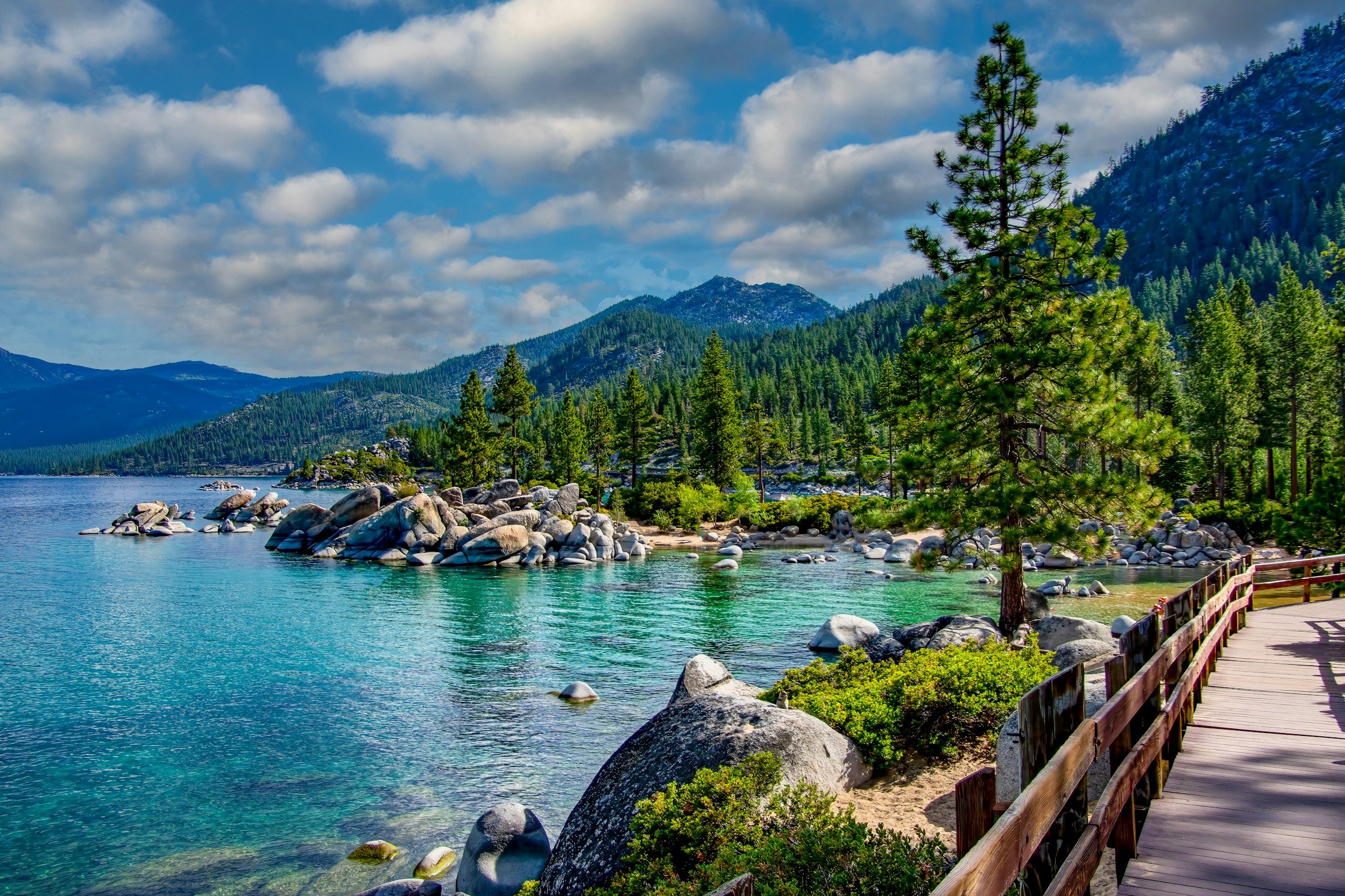 A scenic lakeside view with clear blue water, large rocks along the shore, pine trees, a wooden pathway with a railing, and mountains covered in forests under a partly cloudy sky.