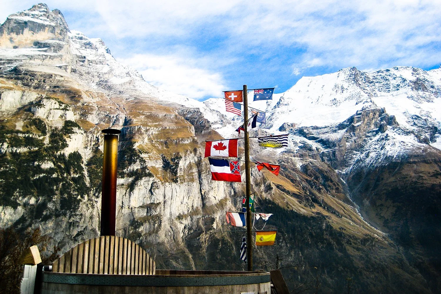 A mountain landscape with snow-capped peaks under a blue sky, featuring a wooden pole with multiple international flags, including Canada, the United States, Switzerland, and others.