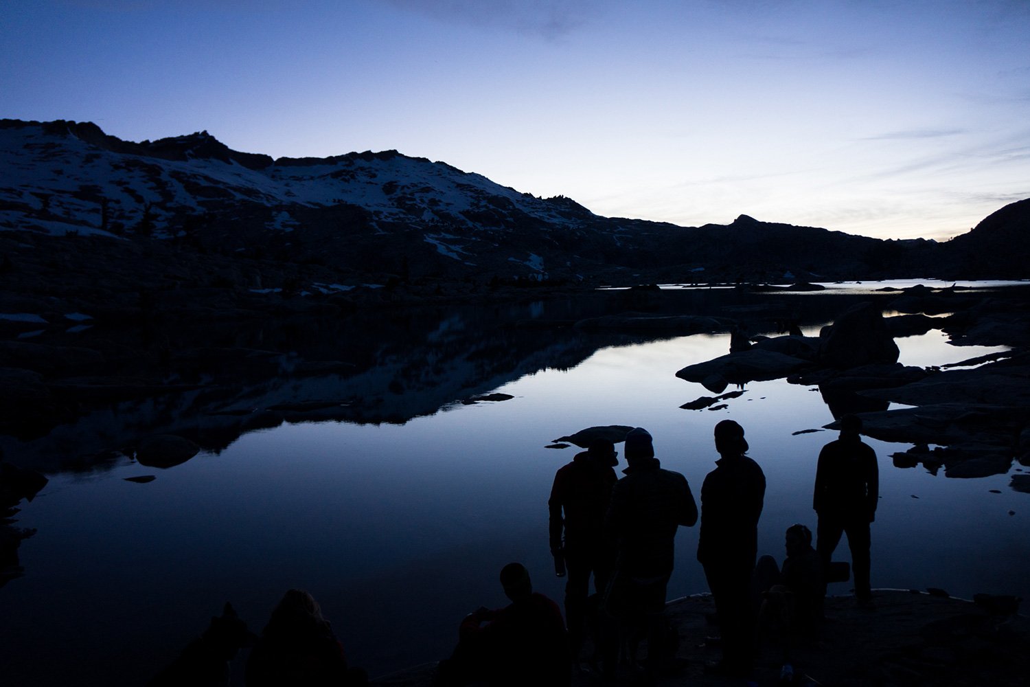 Silhouettes of people near a lake at dusk with snow-covered mountains in the background.