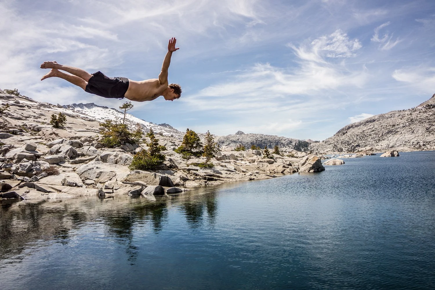 A man diving into a mountain lake surrounded by rocky terrain and sparse trees, under a partly cloudy sky.