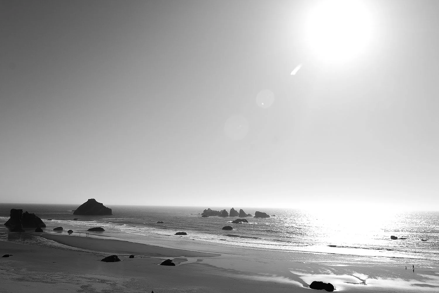 Black and white photo of a beach with large rocks in the water, a sandy shoreline, and the sun shining brightly in the sky.