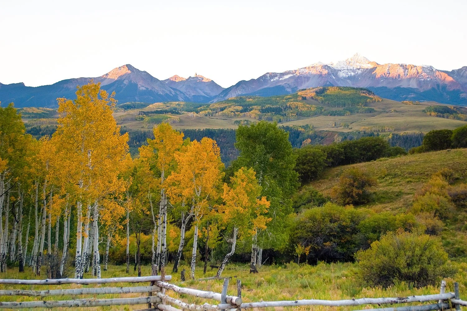 Scenic landscape of mountains with snow caps, rolling green hills, and trees with yellow and green leaves in a fenced field.