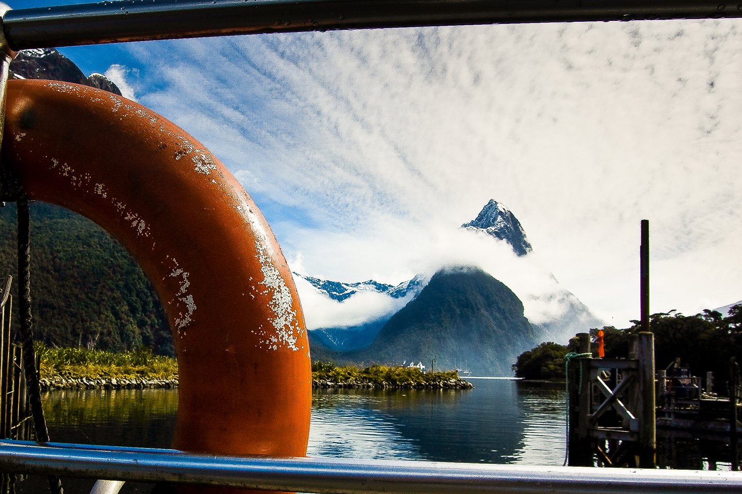 Mountain landscape with steam-covered peak, surrounded by greenery and water, seen from a boat dock with a safety ring and dock structures in the foreground.