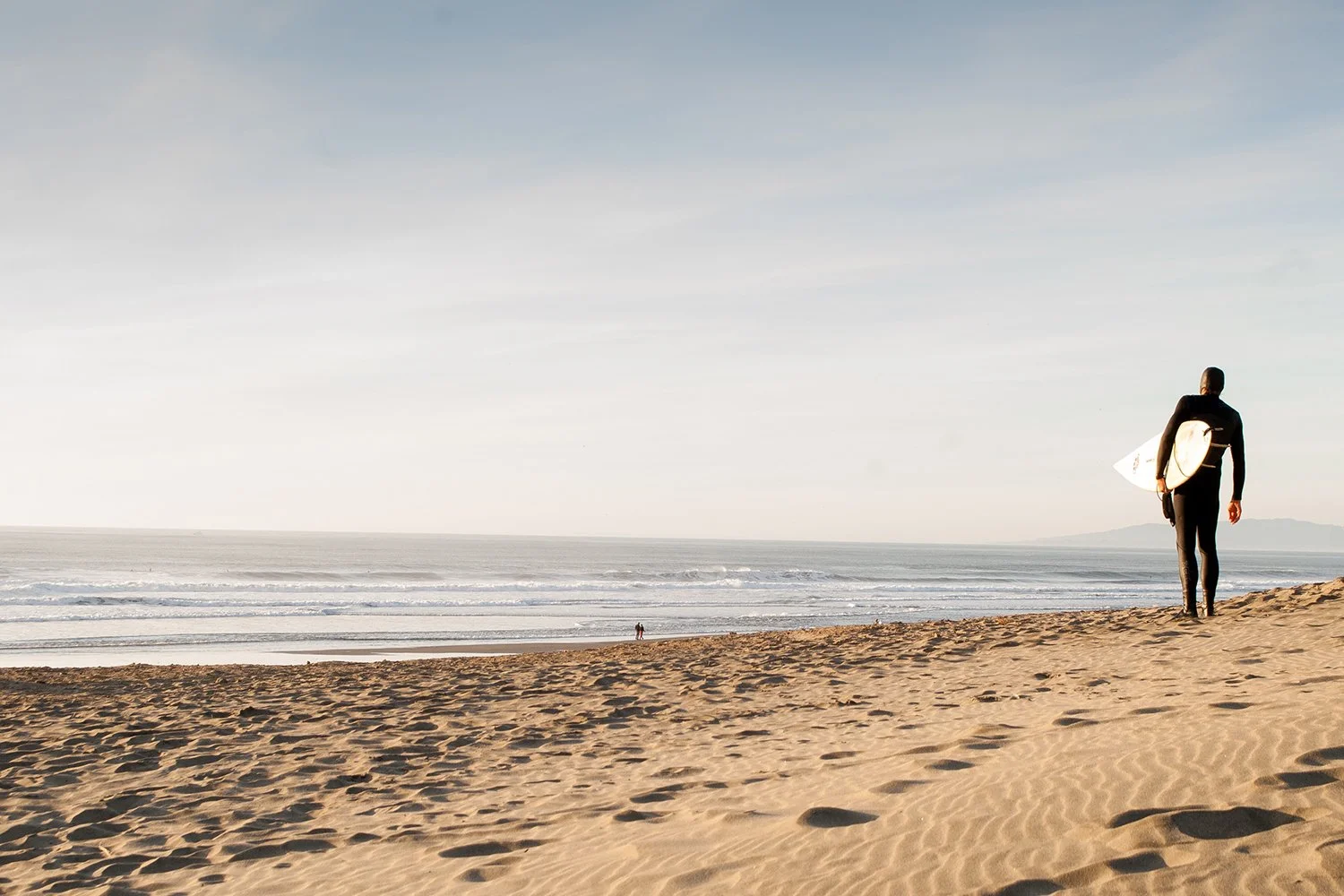 A person in a wetsuit holding a surfboard, walking along the sandy beach towards the ocean, with the sky and distant horizon in view.
