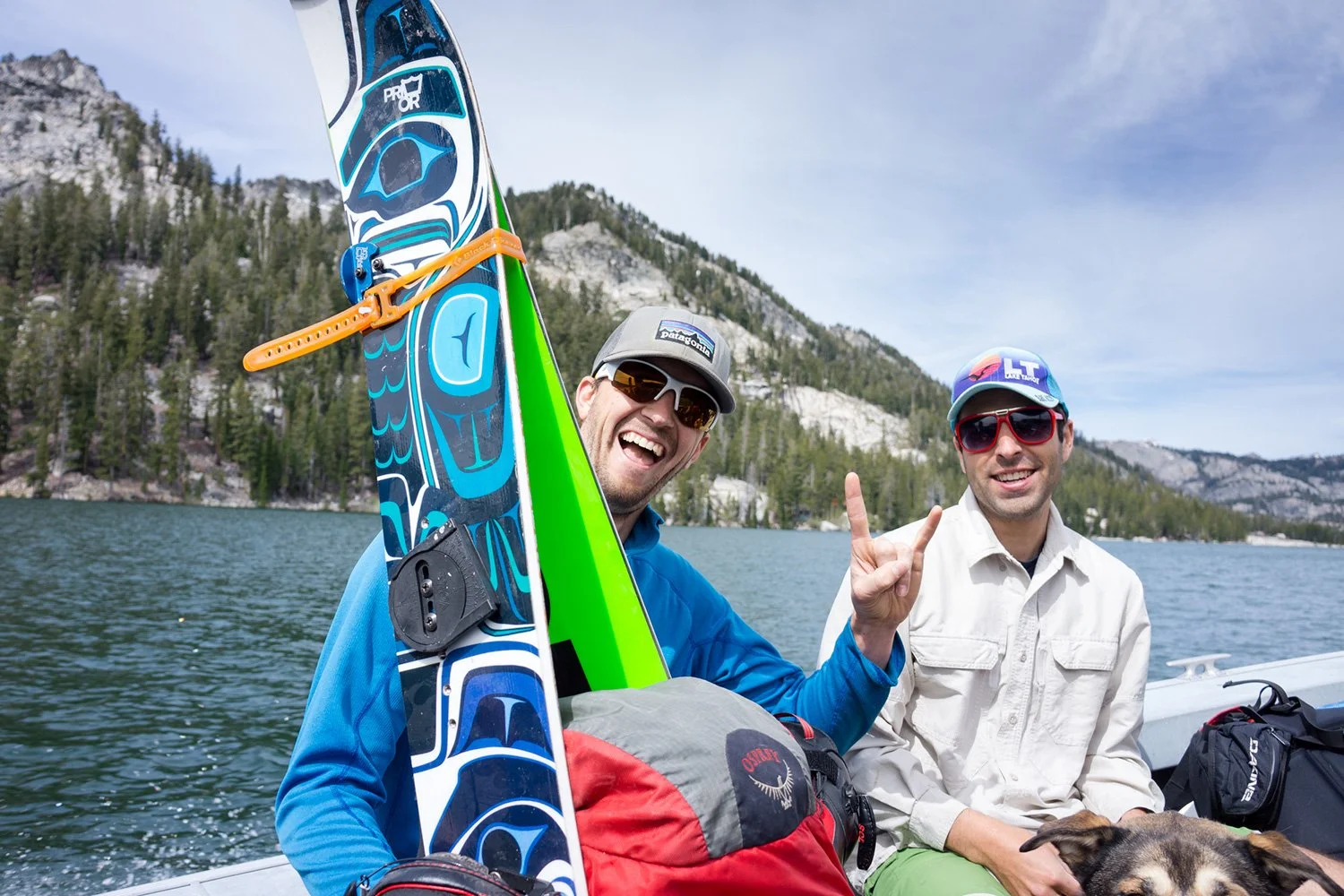Two smiling men on a boat with backpacks and gear, one holding a colorful paddleboard, in a lake surrounded by forested mountains.