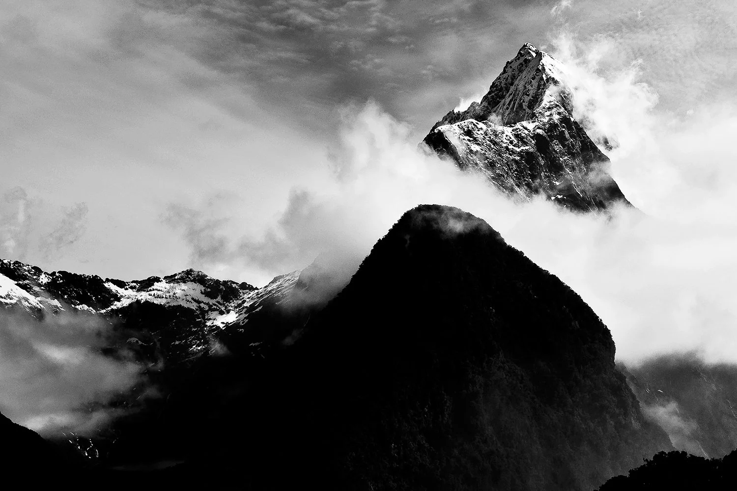 Black and white photo of a mountain range with a tall, snow-capped peak partially obscured by clouds, and smaller forested mountains in the foreground.