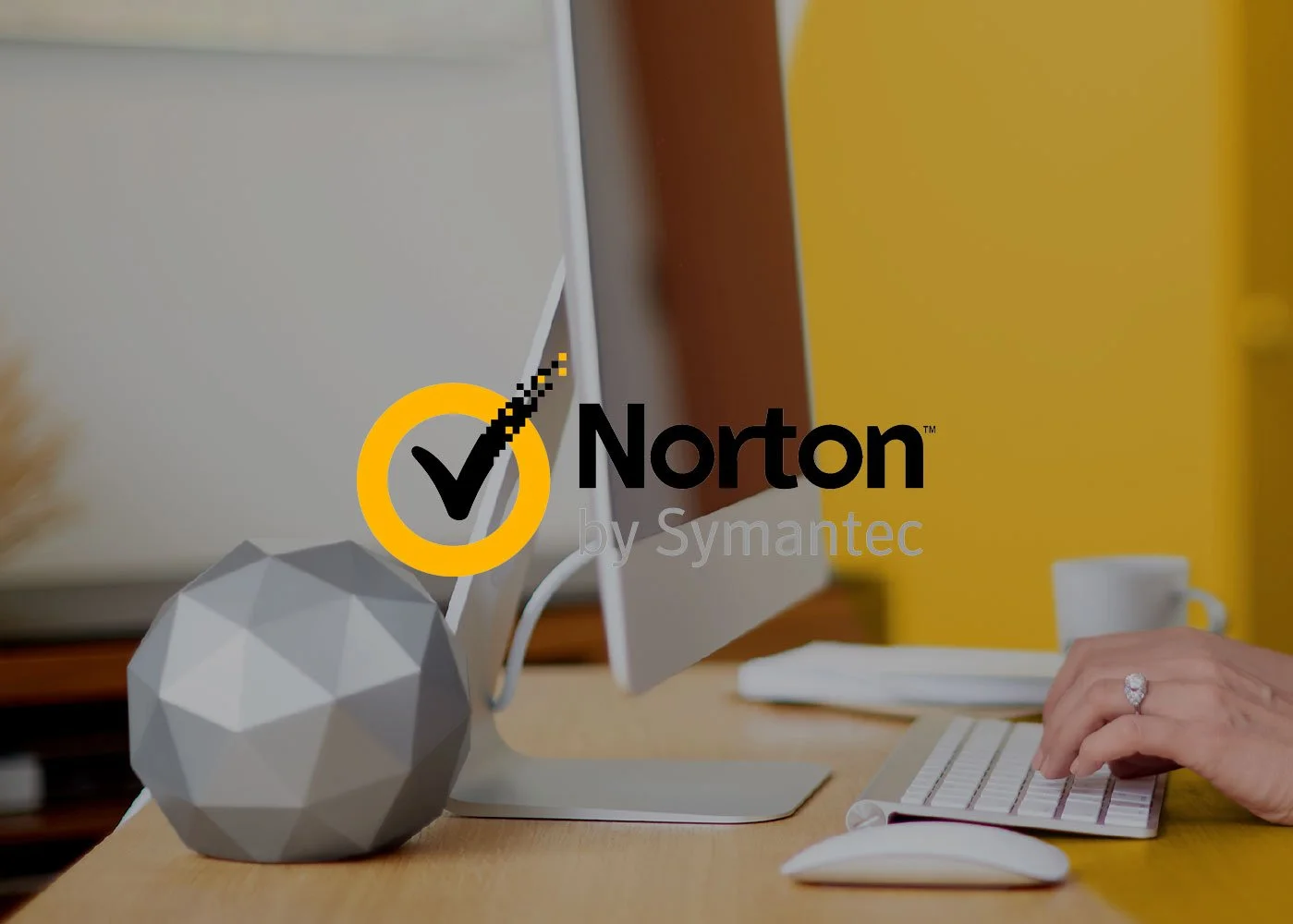 Computer monitor, geometric gray decorative object, keyboard, mouse on a wooden desk in an office setting with a yellow wall and a person's hand with a ring near the keyboard. Overlay text reads 'Norton by Symantec'.