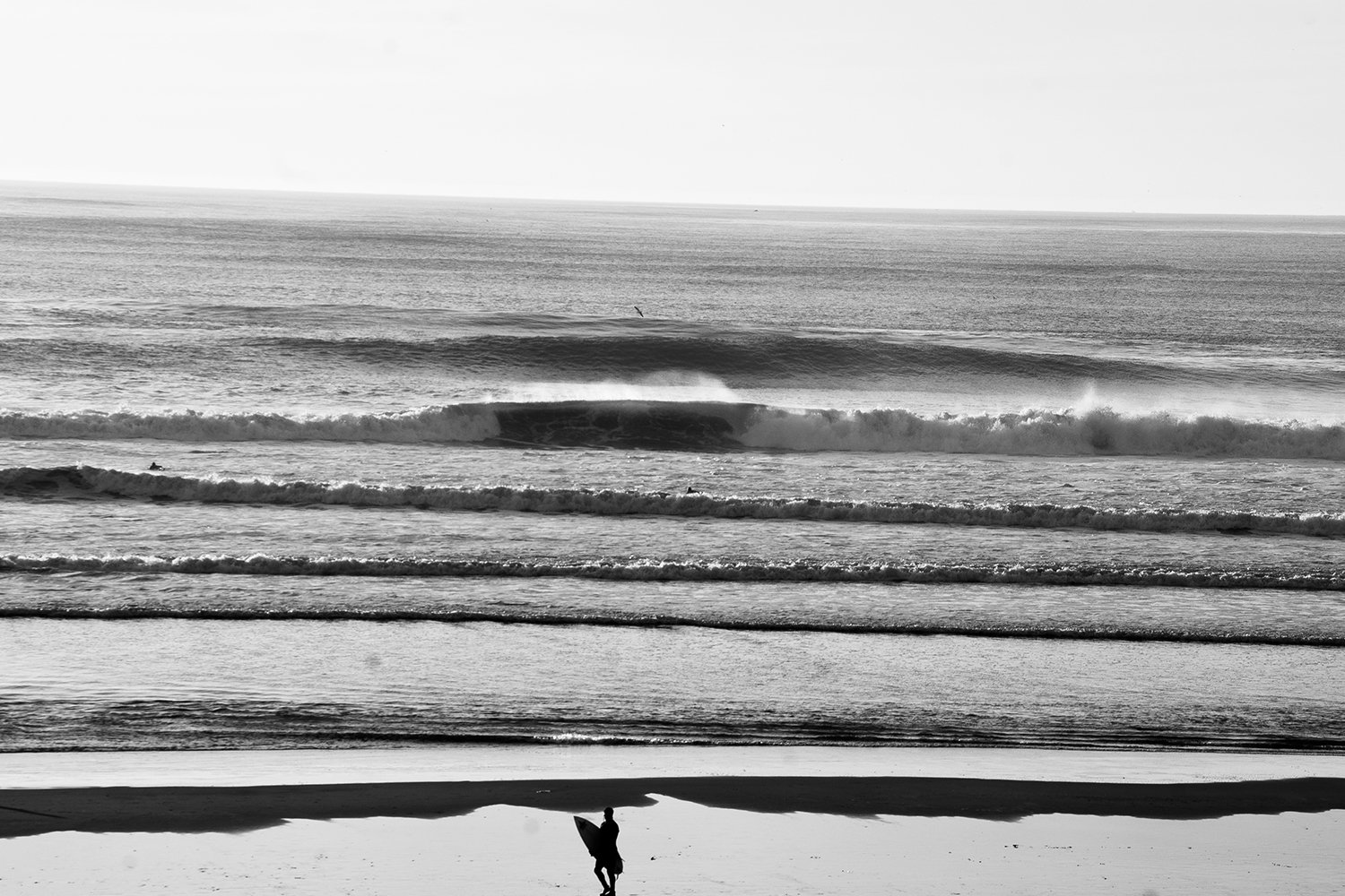 A black and white photo of a person walking on the beach with a surfboard, with waves and the ocean in the background.