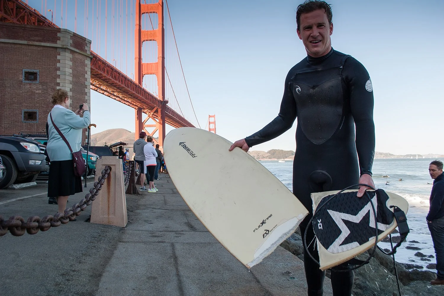 A man in a wetsuit holding a surfboard on a beach near the Golden Gate Bridge, with several people and cars in the background and the bridge spanning across the water.