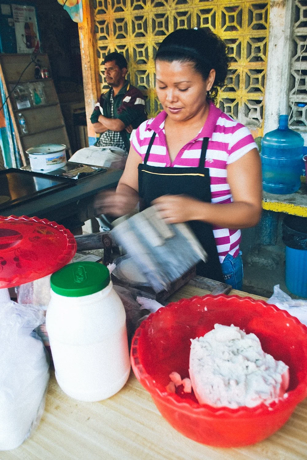 A woman preparing food at a small street stand with a red bowl of dough and a container of white ingredients, standing in front of a yellow lattice wall with a man in the background.