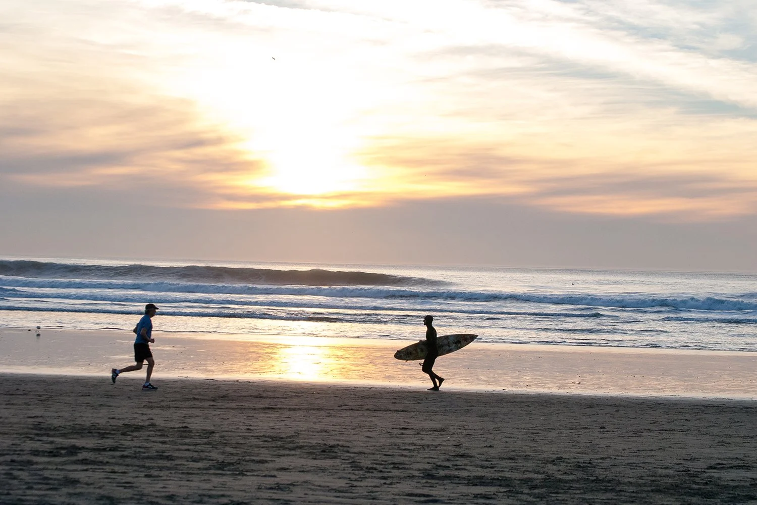 A person with a surfboard walking on the beach at sunset while another person jogs nearby, with ocean waves in the background.