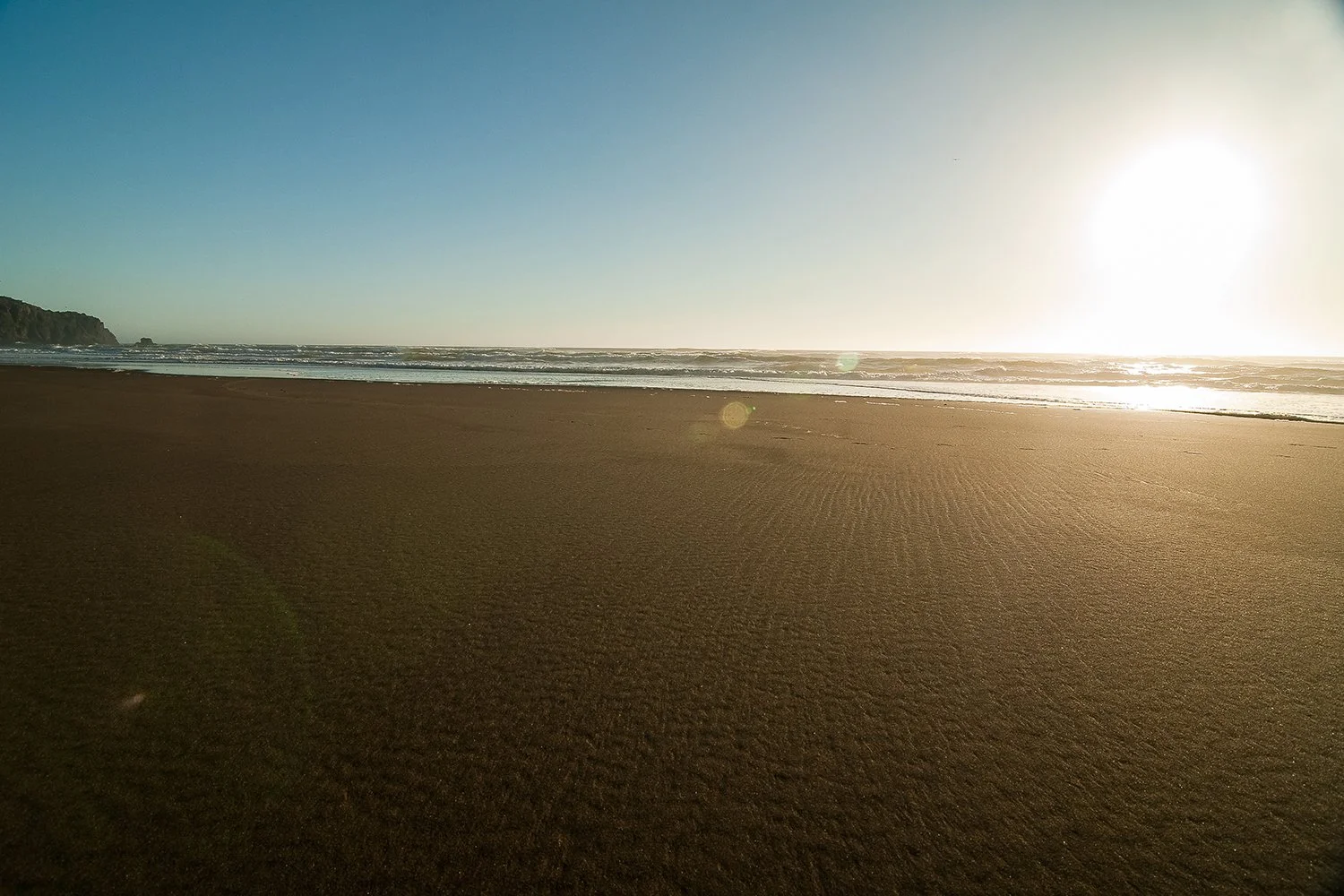 Empty sandy beach with gentle waves and a setting sun on the horizon.