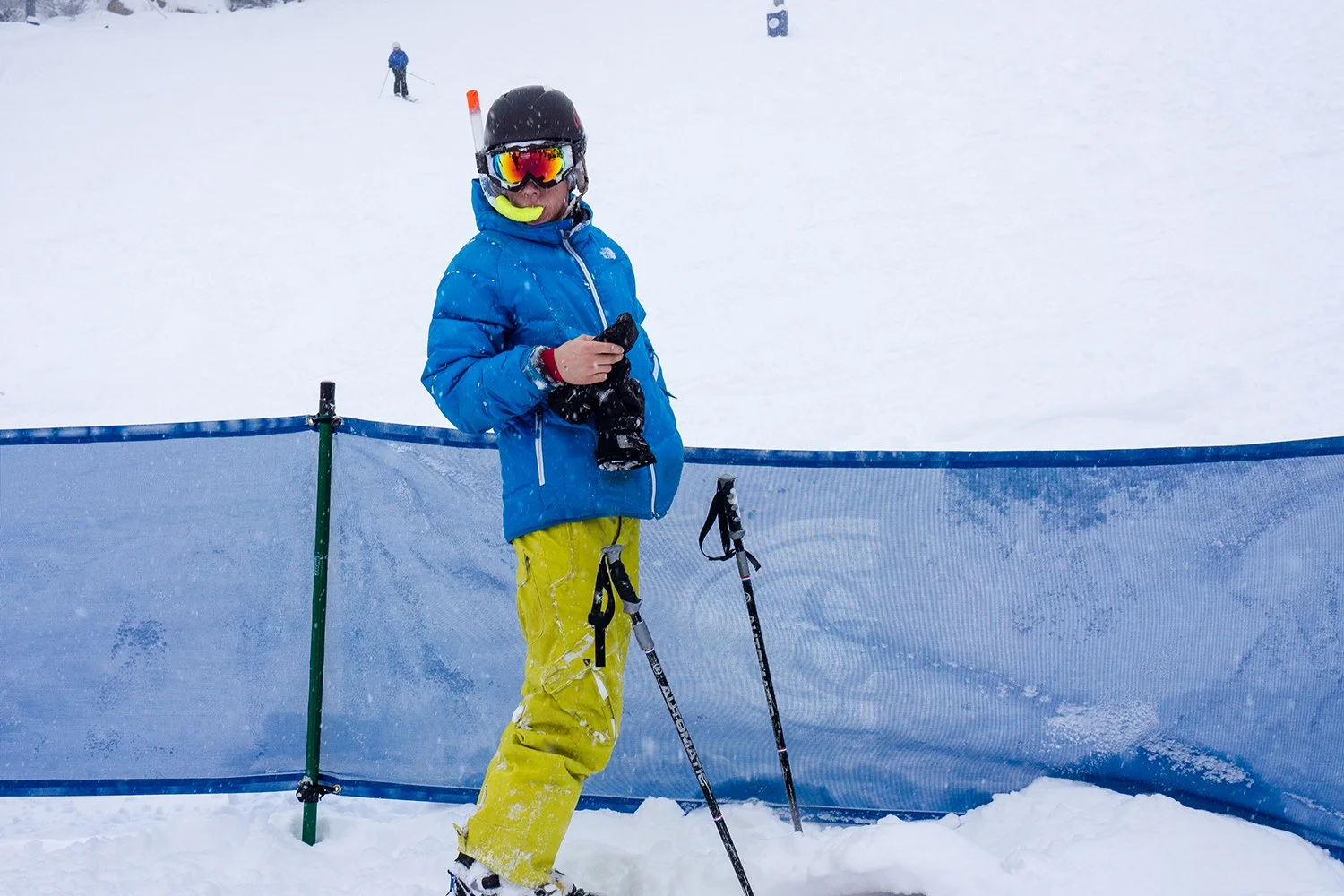 A person standing in snowy ski area wearing a black helmet, ski goggles, a blue jacket, and yellow ski pants, holding a phone, with ski poles on the snow and a blue safety net behind them.