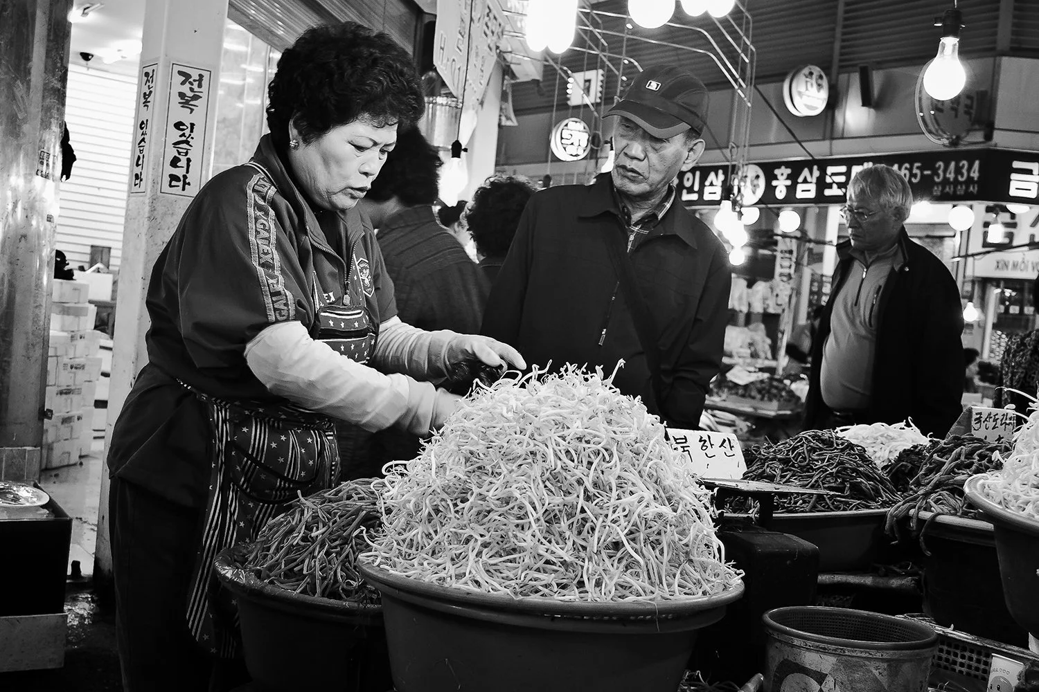 Black and white photo of a woman shopping at an Asian market, with three other people nearby, looking at a large pile of bean sprouts on a market stall.