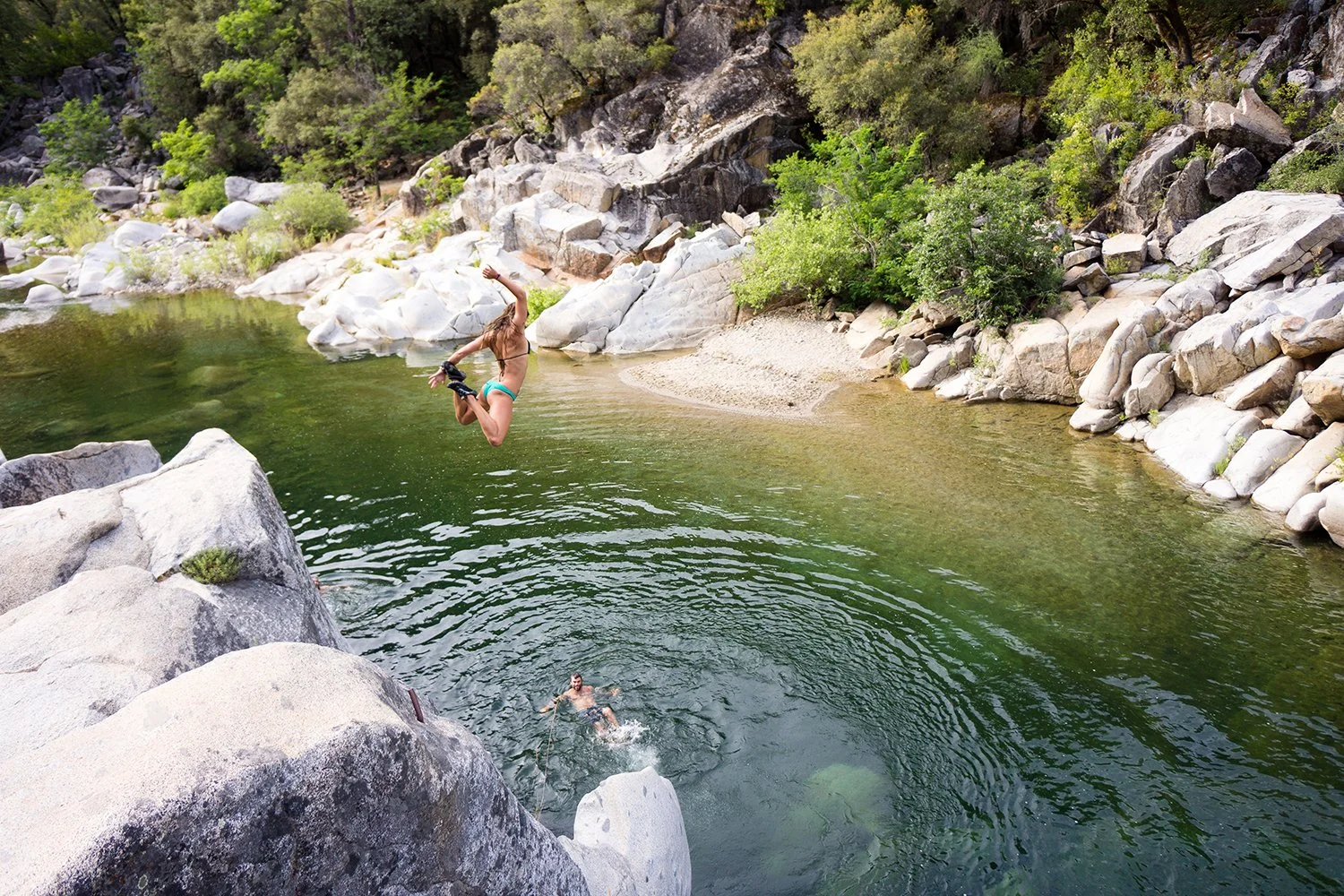 A person jumping off a large rock into a green river in a rocky, forested area.