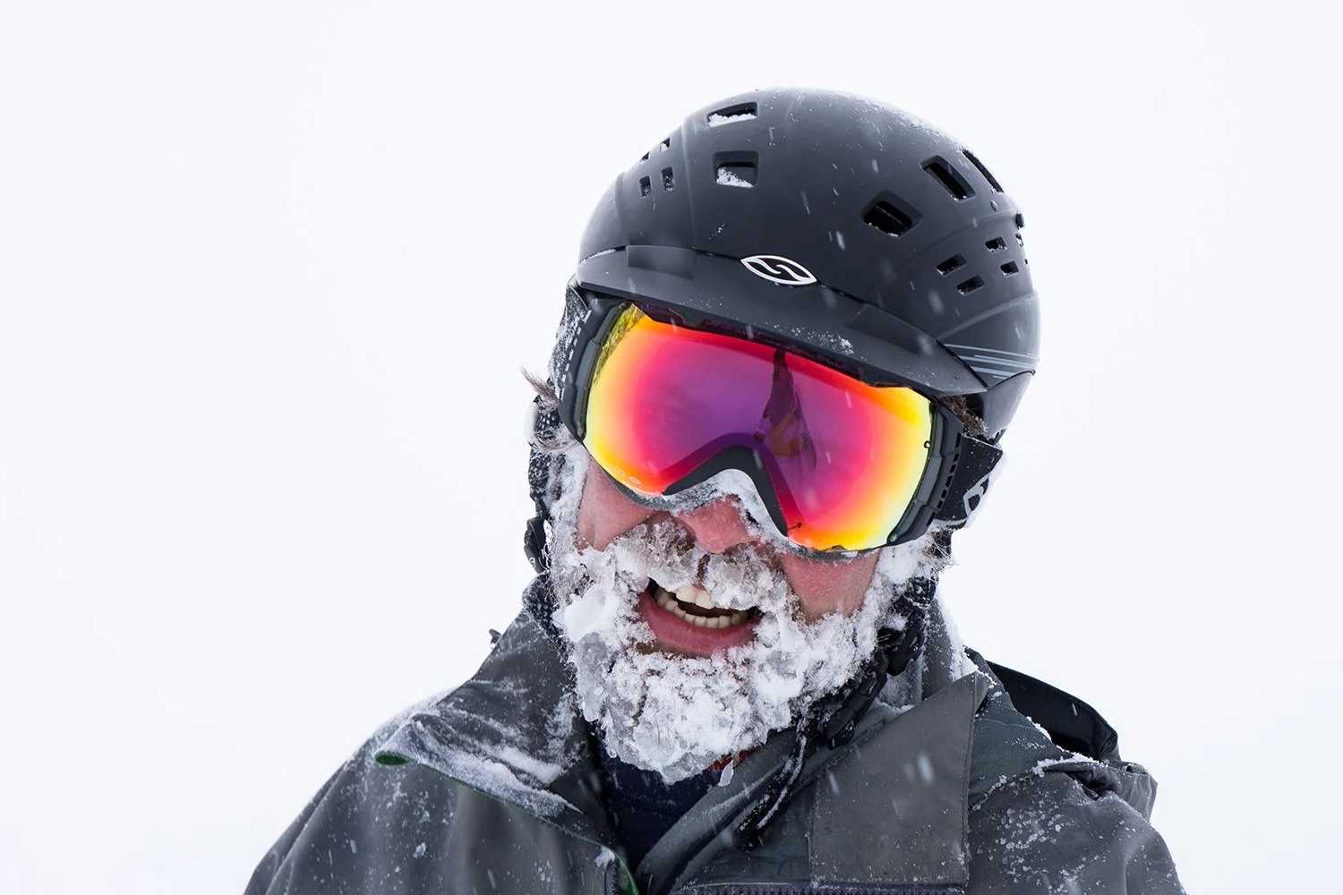 Smiling man in ski gear with a beard, wearing a black helmet, reflective ski goggles, and a gray jacket, outdoors in snowy weather.